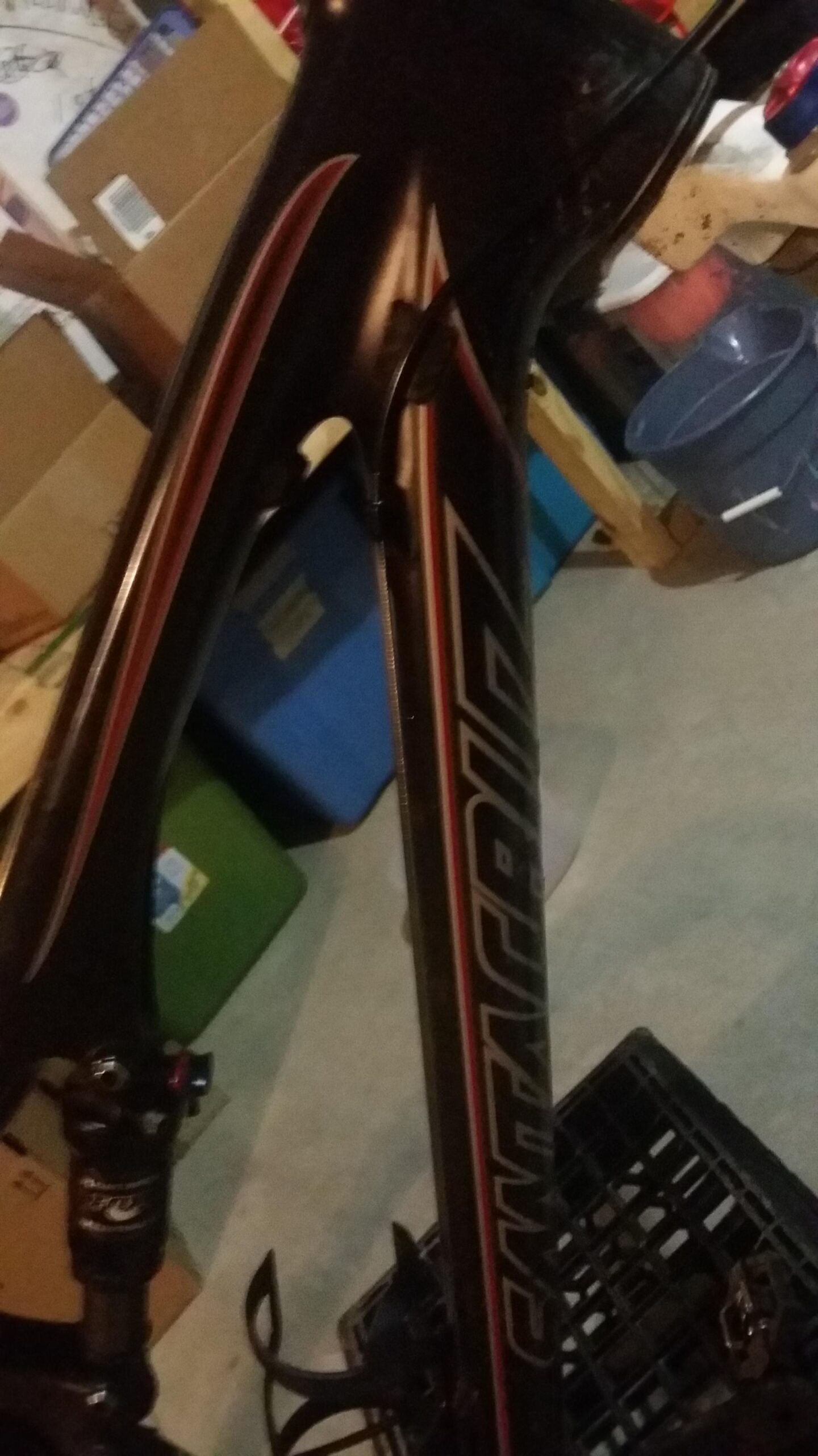 Santa Cruz Tallboy Carbon: A close-up image of a mountain bike frame with prominent branding, featuring a dark finish with red accents. The bike is positioned in a cluttered garage environment, with boxes and bins visible in the background. The shot captures the frame's design details and the rear shock absorber.
