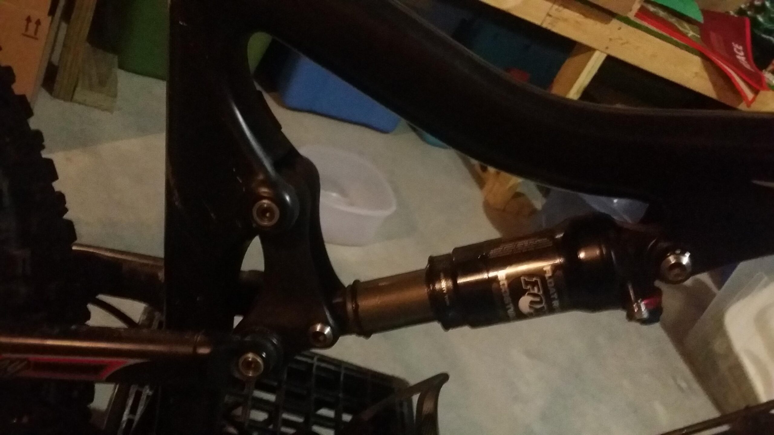 Santa Cruz Tallboy Carbon: Close-up view of a mountain bike’s rear suspension system, featuring the shock absorber and connection points on the frame. The setting appears to be a garage, with various storage items blurred in the background.