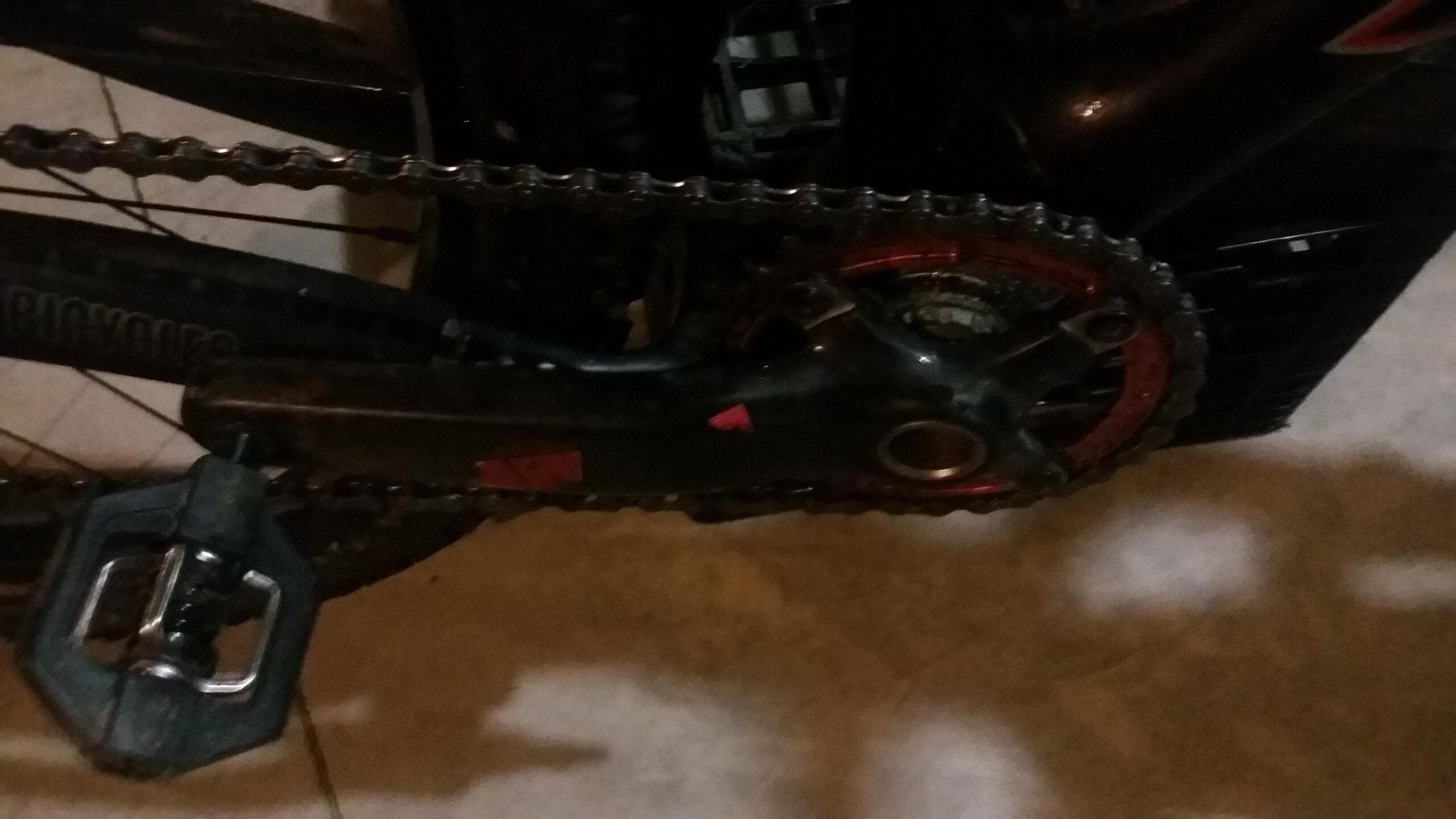 Santa Cruz Tallboy Carbon: Close-up of a bicycle's crankset and chain, featuring a black and red gear combination, with a pedal visible on the left side. The background shows a tiled floor, indicating an indoor setting.