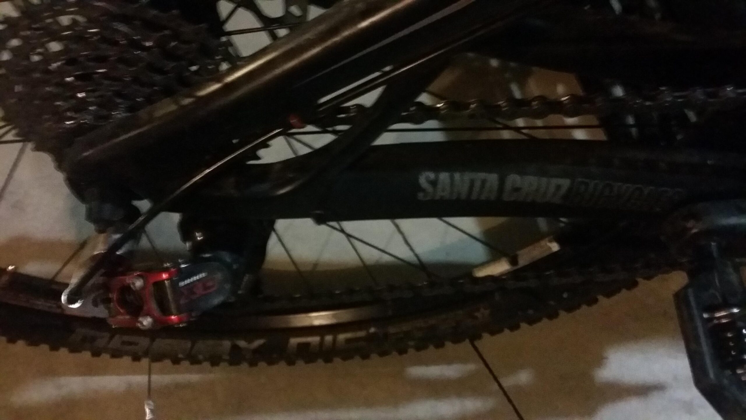 Santa Cruz Tallboy Carbon: Close-up image of a black mountain bike's rear drivetrain, featuring a visible chain, cassette, and a red rear derailleur. The bike frame is labeled with "Santa Cruz Bicycles." The background shows a neutral surface.