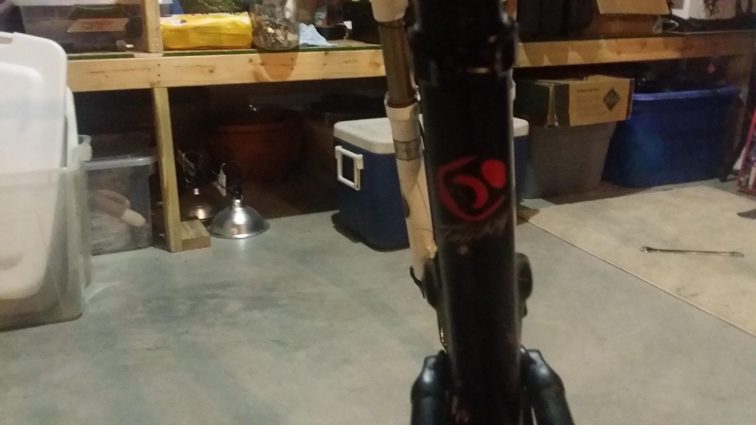 Santa Cruz Tallboy Carbon: A close-up view of a black bicycle frame with a red logo, set against a dimly lit garage background containing various stored items like coolers, boxes, and tools. The garage floor is concrete and shows signs of wear.
