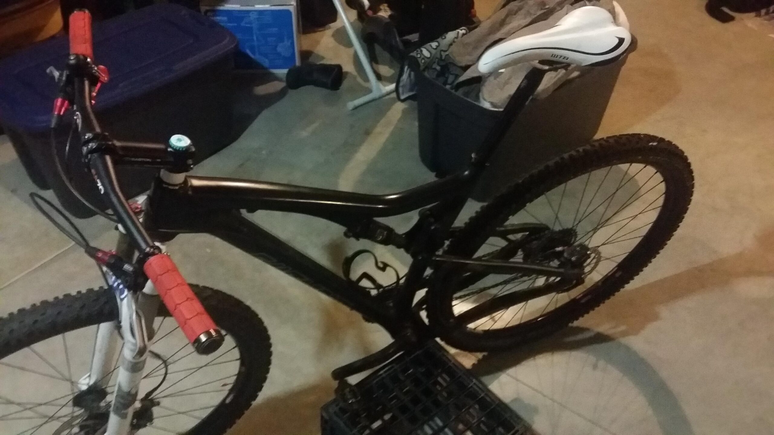 Santa Cruz Tallboy Carbon: A black mountain bike with red handlebar grips and a white saddle, leaning against a black storage crate in a garage setting. The background features various household items and storage containers.