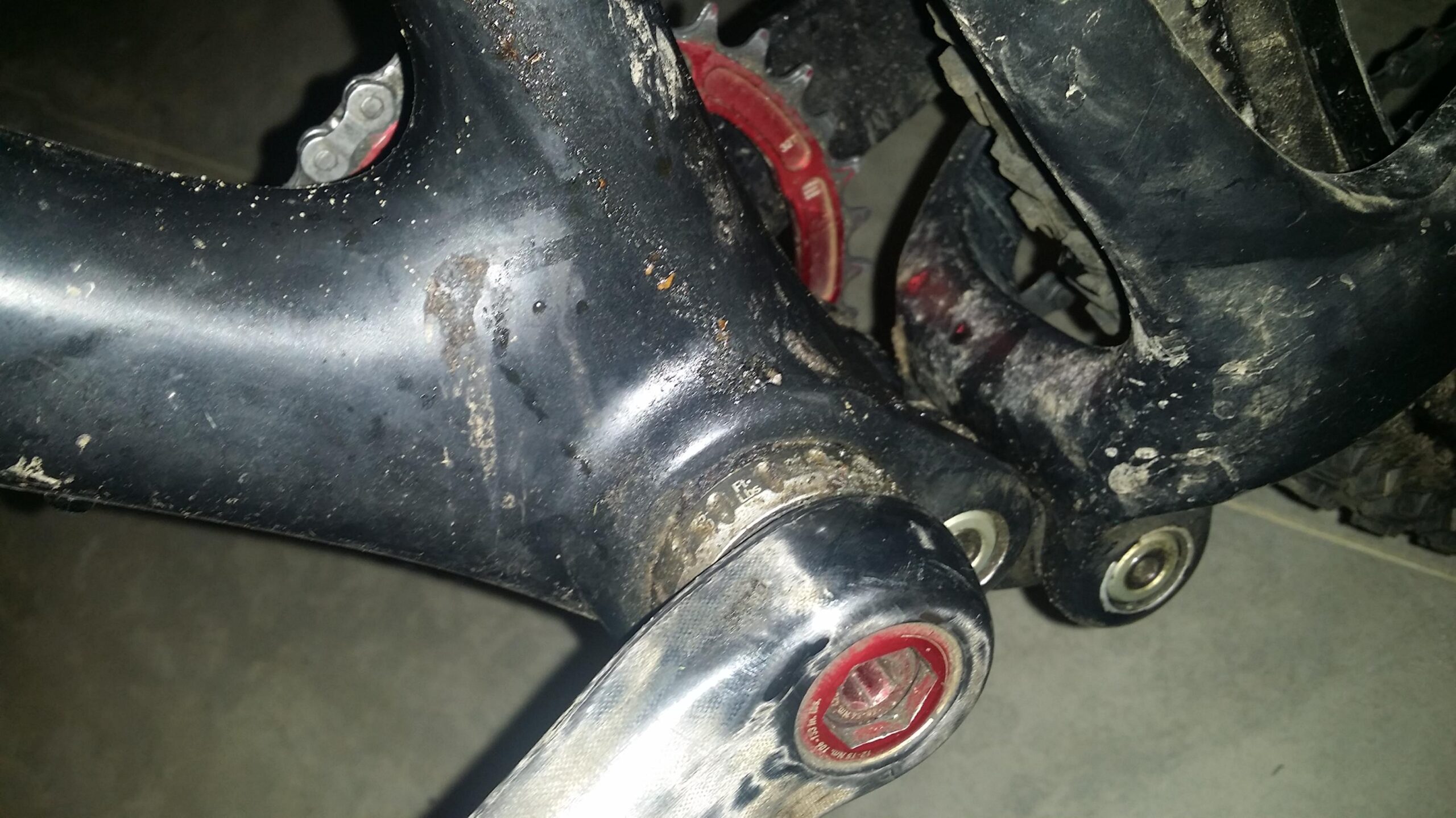 Santa Cruz Tallboy Carbon: Close-up view of a bicycle's bottom bracket area, featuring a black frame with some dirt and wear. The crank arm is visible, showing a metallic finish and red accents, with visible threads and a bearing. The background is blurred, indicating a garage or indoor setting.