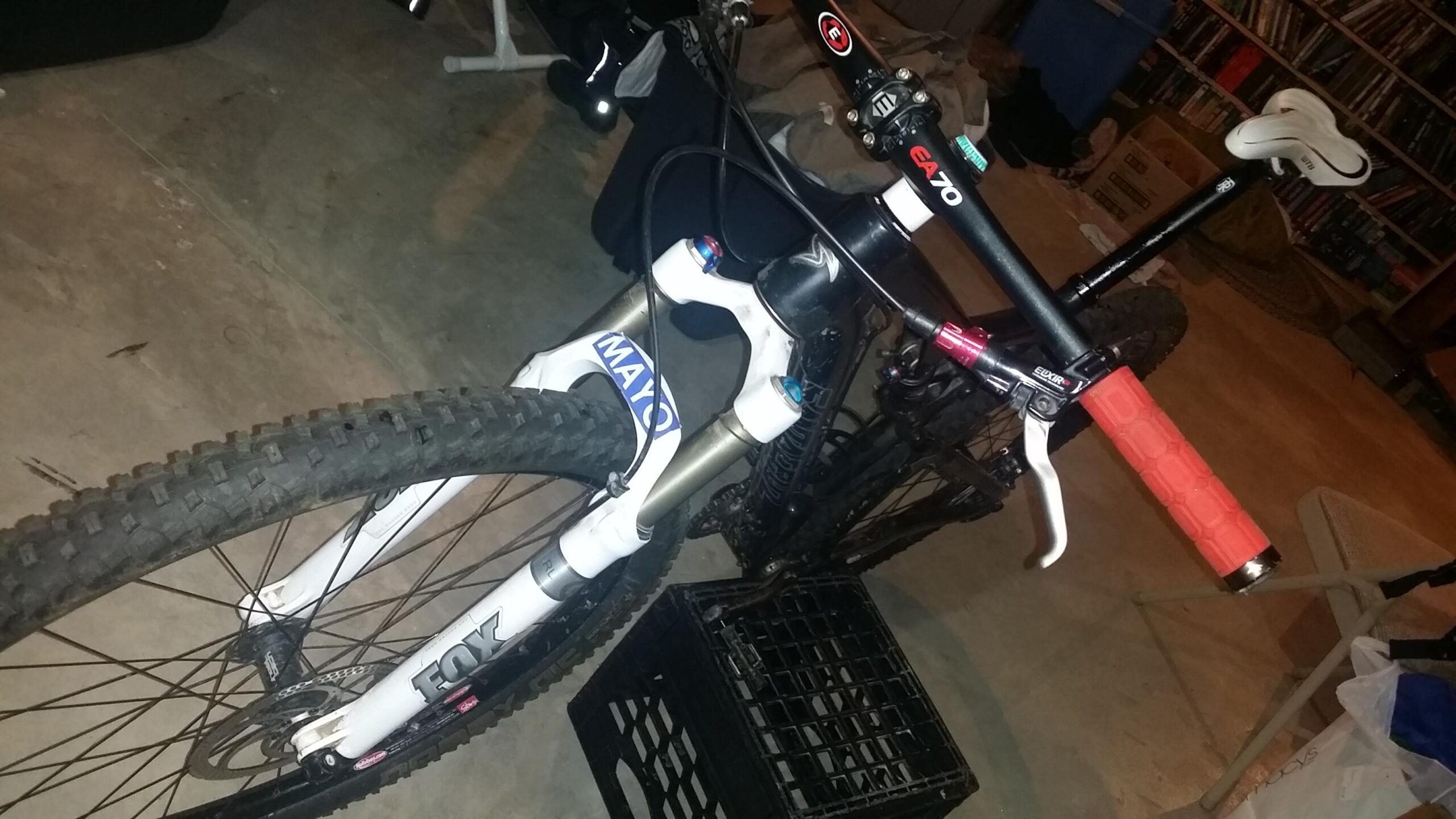 Santa Cruz Tallboy Carbon: A close-up view of a mountain bike featuring a white and black front fork branded with "FOX," a black handlebar with red grips, and a saddle in the background. The bike is positioned on a concrete floor, along with some clutter visible in the background.
