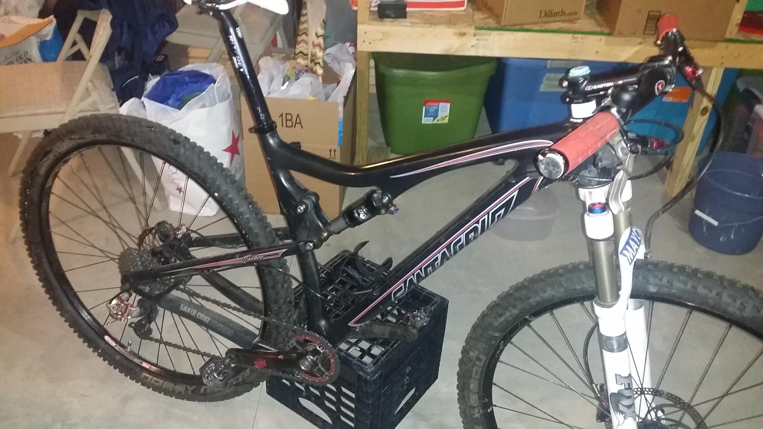Santa Cruz Tallboy Carbon: A mountain bike with a black frame and red accents is positioned on a crate in a cluttered garage. The bike features thick, knobby tires and a white front fork with branding. Various items, including bags and boxes, are visible in the background, indicating a storage area.