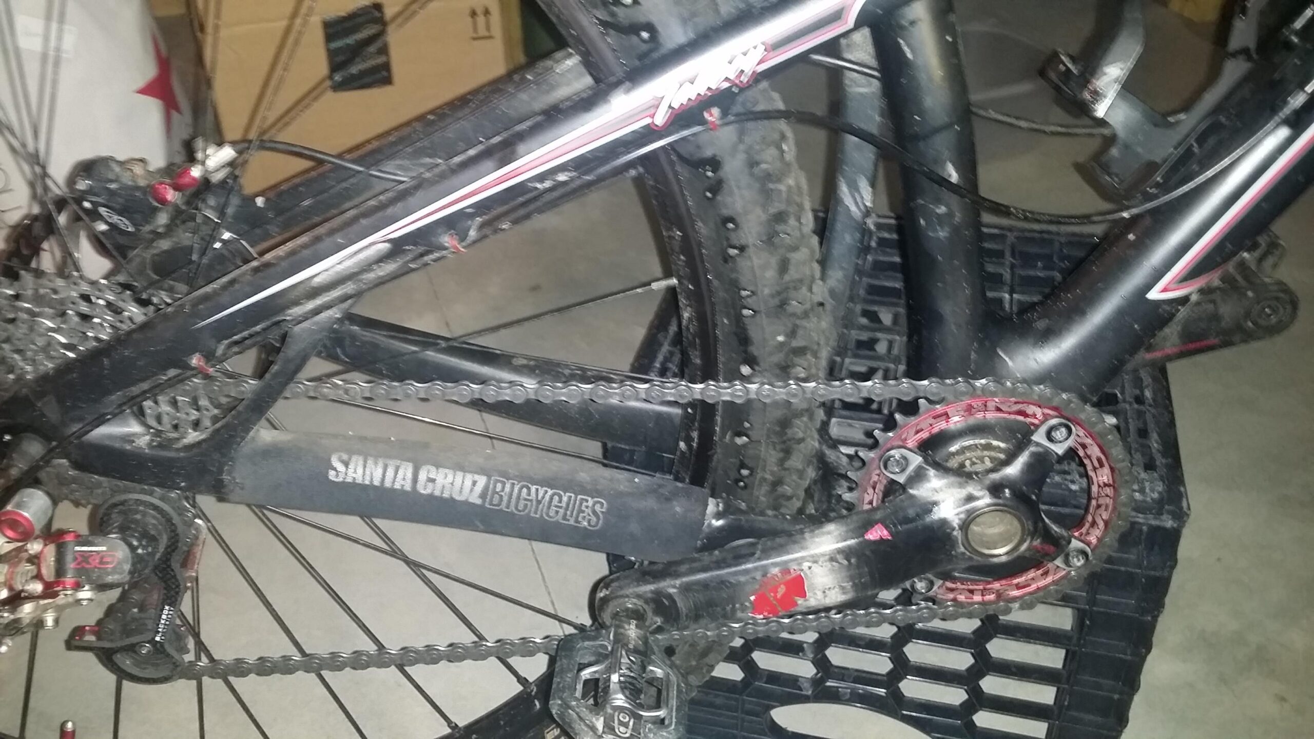 Santa Cruz Tallboy Carbon: A close-up image of a mountain bike with a Santa Cruz Bicycles frame, showing a dirt-covered chain, pedals, and rear derailleur. The bike is positioned on a black plastic crate, with a blurred background of boxes and a floor. The image highlights the components and condition of the bike.
