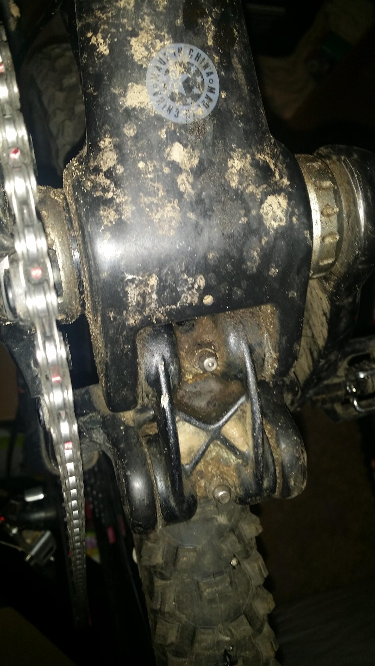 Santa Cruz Tallboy Carbon: Close-up view of a mountain bike's rear suspension link, showing dirt and grime, with the chain and tire partially visible in the background.