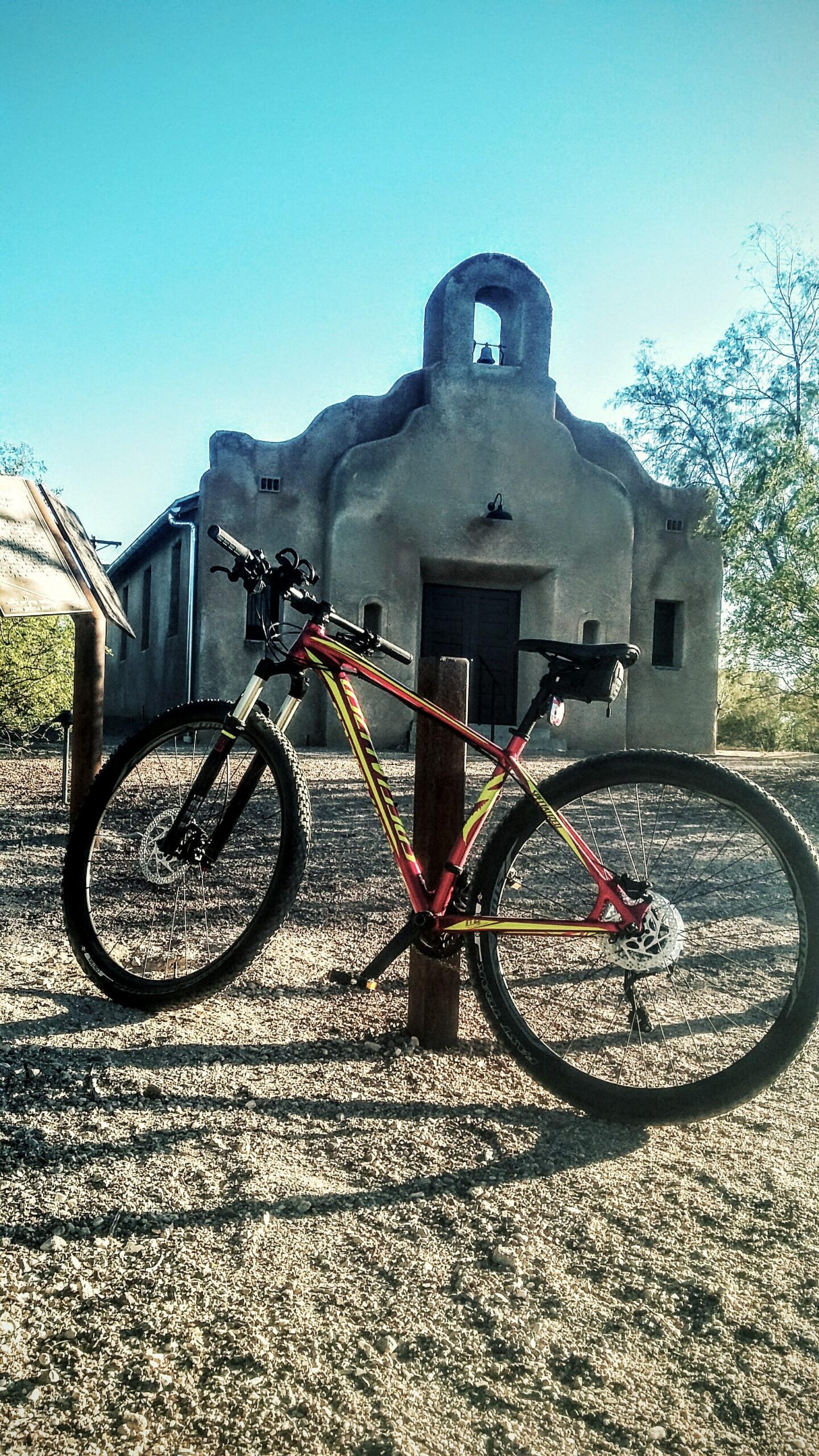 Specialized Crave Pro 29: A mountain bike leaning against a wooden post in front of a rustic adobe building with a bell tower. The scene is set under a clear blue sky, with sunlight casting shadows on the gravel ground.