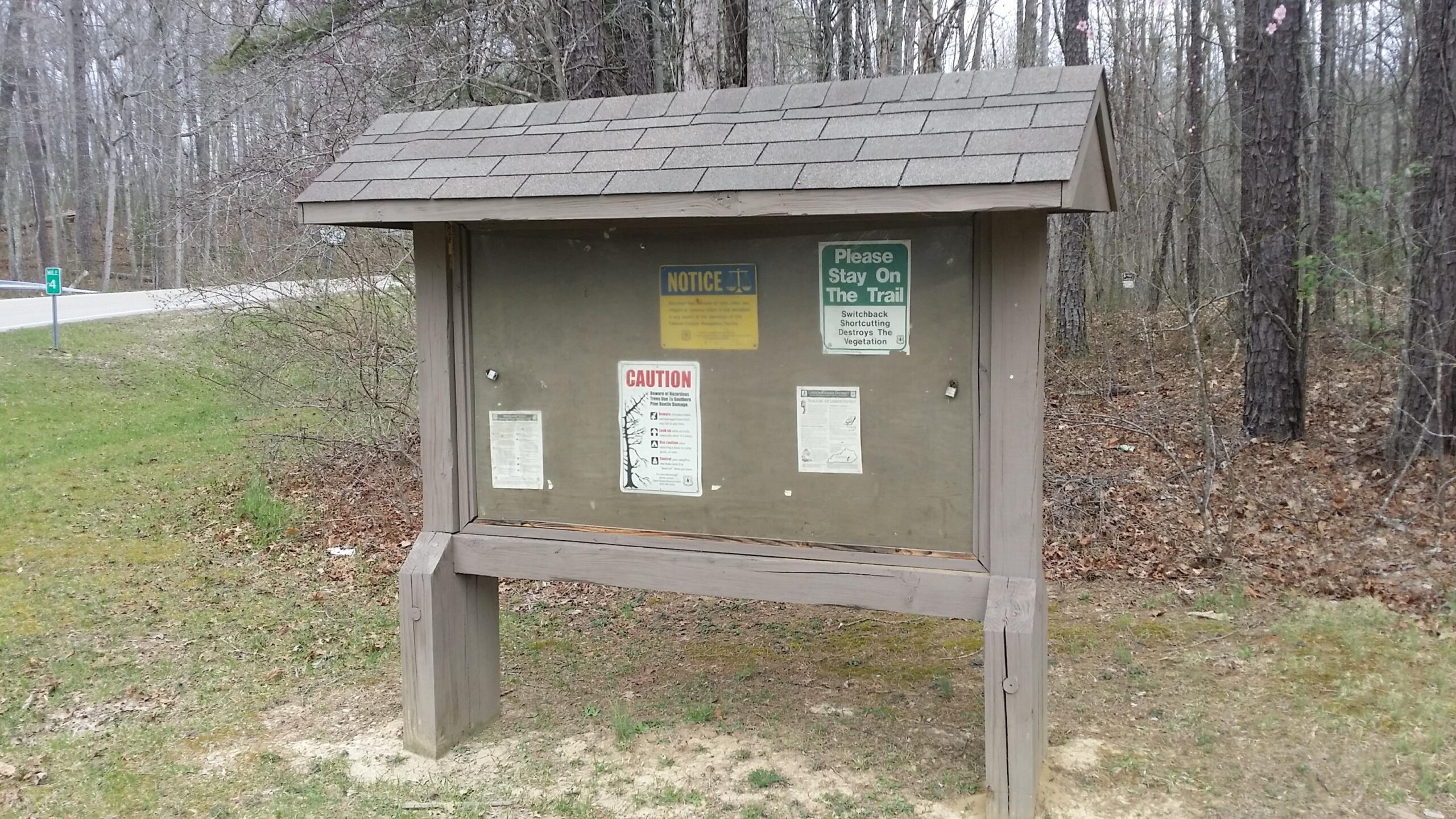 A wooden informational signboard with a shingled roof, displaying several notices and warnings about trail usage. The background features a wooded area with bare trees, and a gravel path can be seen in the distance. A road sign indicating a trail marker is visible on the left side of the image. Sheltowee Trace - Laurel Lake Trail mountain bike trail.