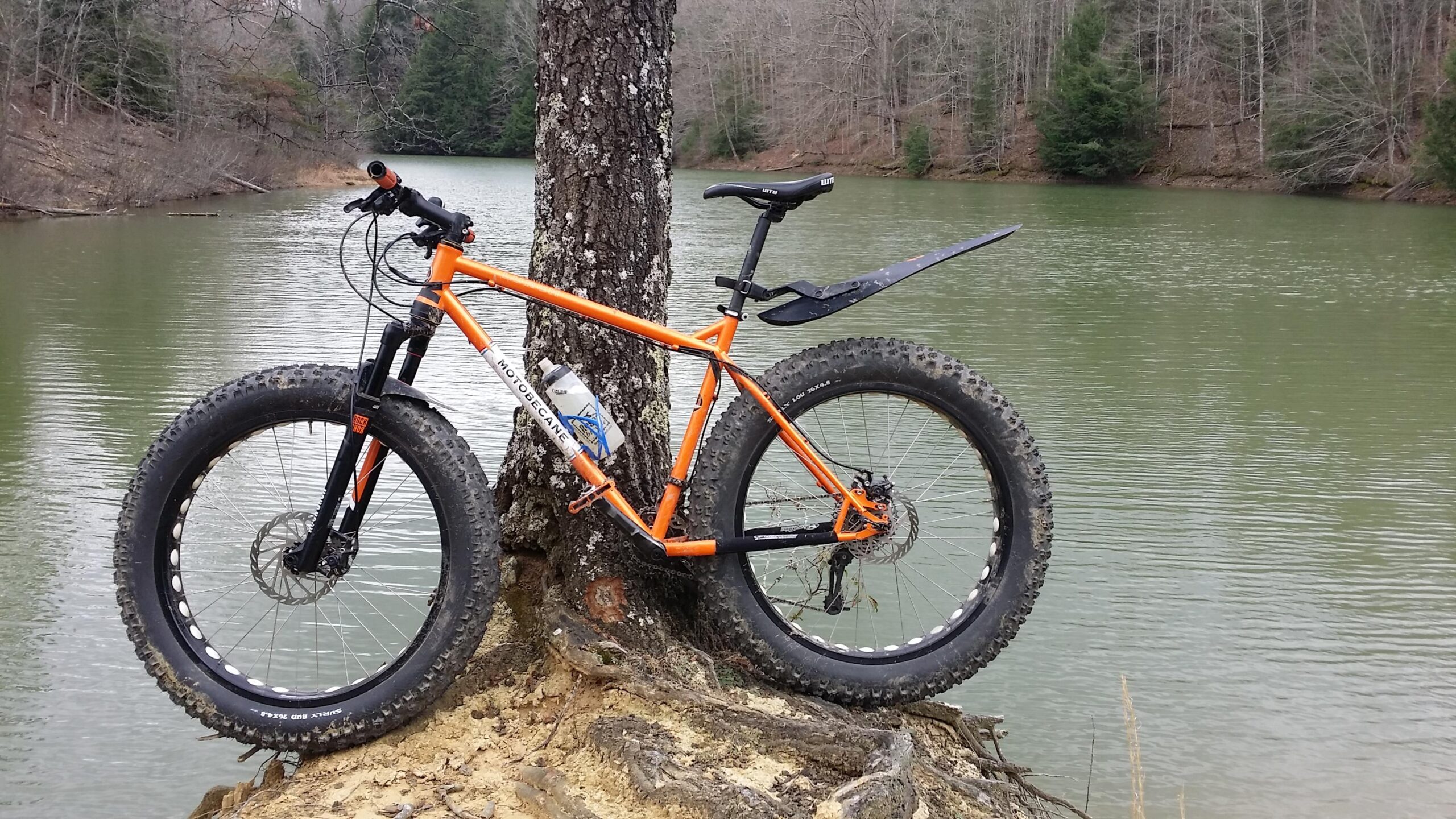 A fat bike with an orange frame is leaning against a tree near a calm, greenish lake. The bike has wide tires, a water bottle mounted on the frame, and the background features sparse trees without leaves, indicating a possibly cool season. Sheltowee Trace - Laurel Lake Trail mountain bike trail.
