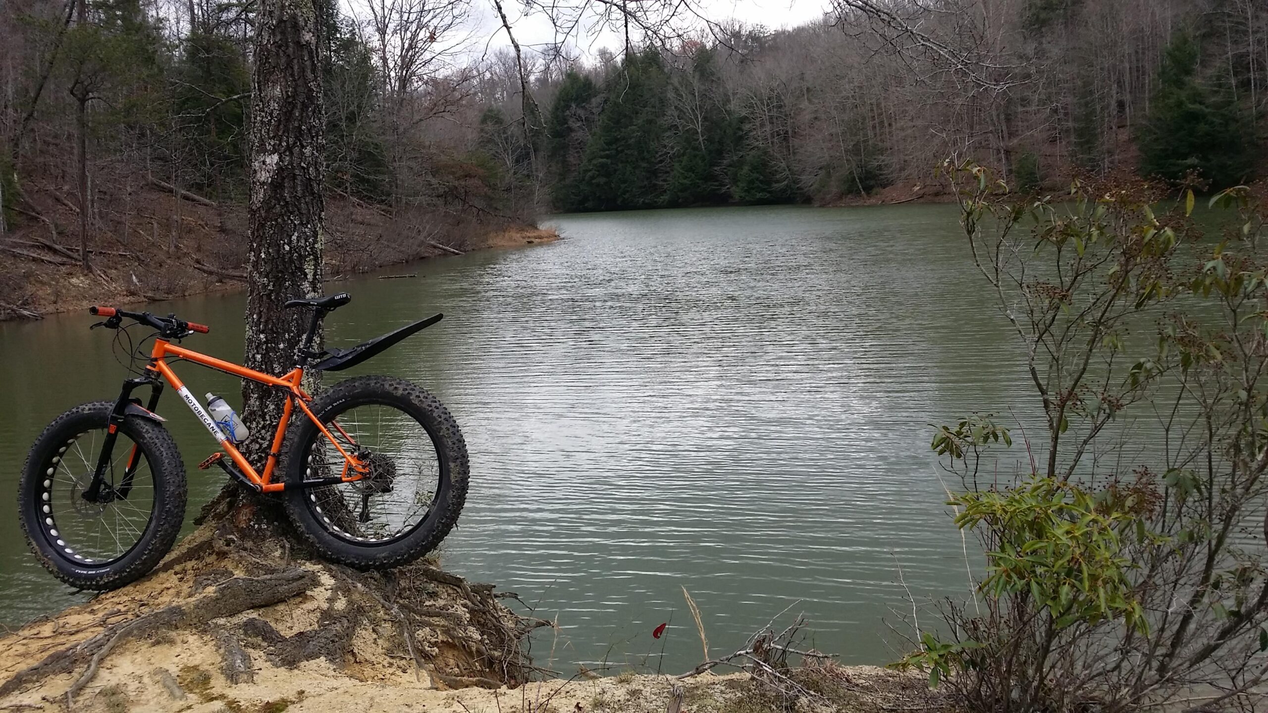 A bright orange fat bike is parked next to a calm lake, surrounded by trees. The bike is positioned on a sandy bank, partially obscured by tree roots, with a water bottle mounted on its frame. The scenery features a mix of bare trees and evergreens in the background, with a cloudy sky overhead. Sheltowee Trace - Laurel Lake Trail mountain bike trail.