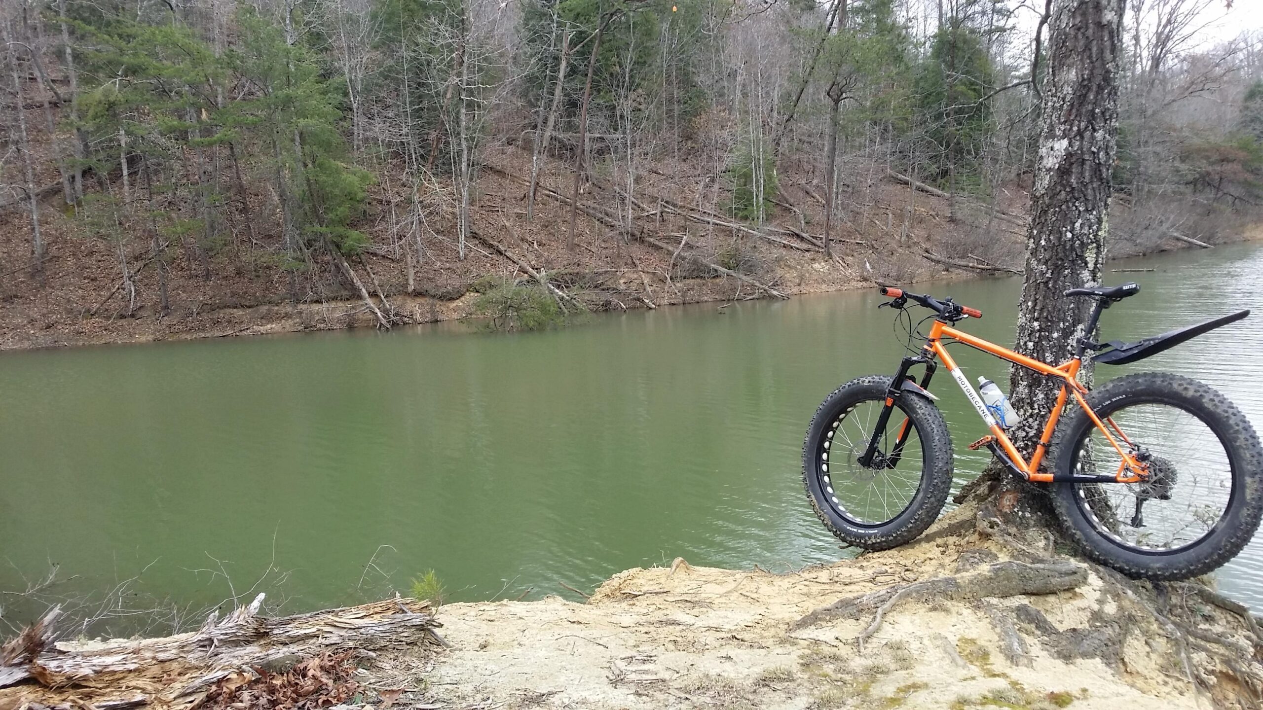 A bright orange mountain bike with fat tires rests against a tree by a calm, green-tinted lake. The background features a wooded area with leafless trees and rocky terrain, suggesting an autumn or early spring setting. Sheltowee Trace - Laurel Lake Trail mountain bike trail.