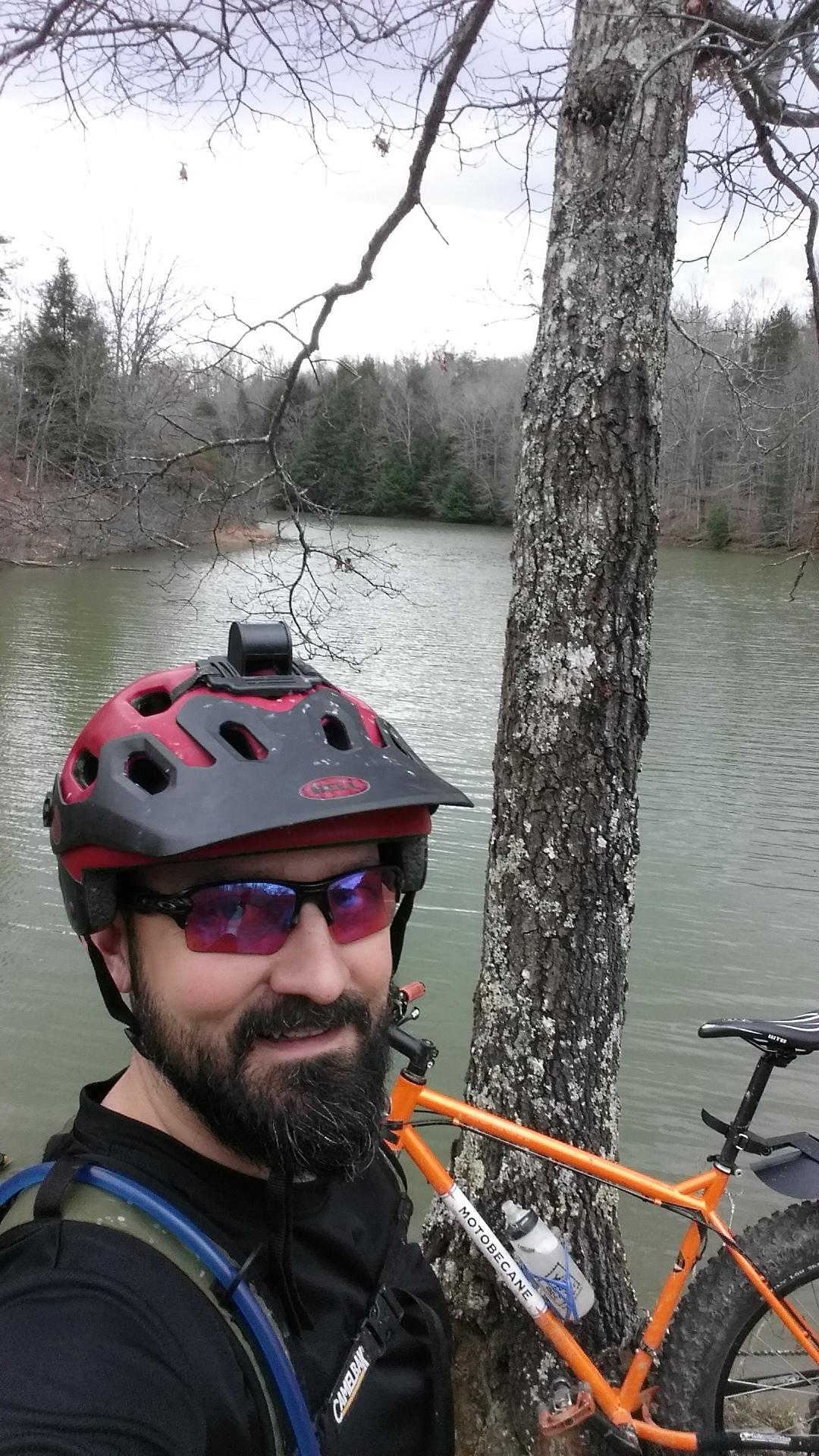 A person wearing a red biking helmet and sunglasses stands by a tree near a lake, smiling for the camera. An orange mountain bike is leaning against the tree, with a water bottle attached. The background features a calm lake and sparse trees, indicating a natural outdoor setting. Sheltowee Trace - Laurel Lake Trail mountain bike trail.