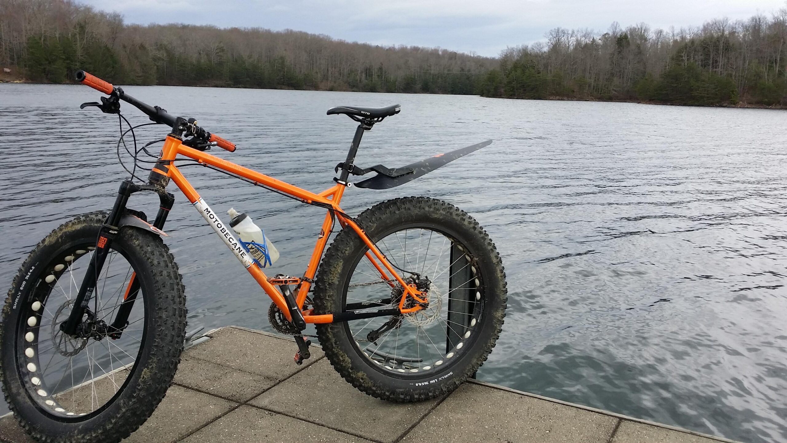 A bright orange fat bike with wide tires is parked on a dock beside a calm lake, surrounded by trees in the background. The bike has a water bottle attached to its frame and features a mudguard. The water appears still, reflecting the overcast sky. Sheltowee Trace - Laurel Lake Trail mountain bike trail.