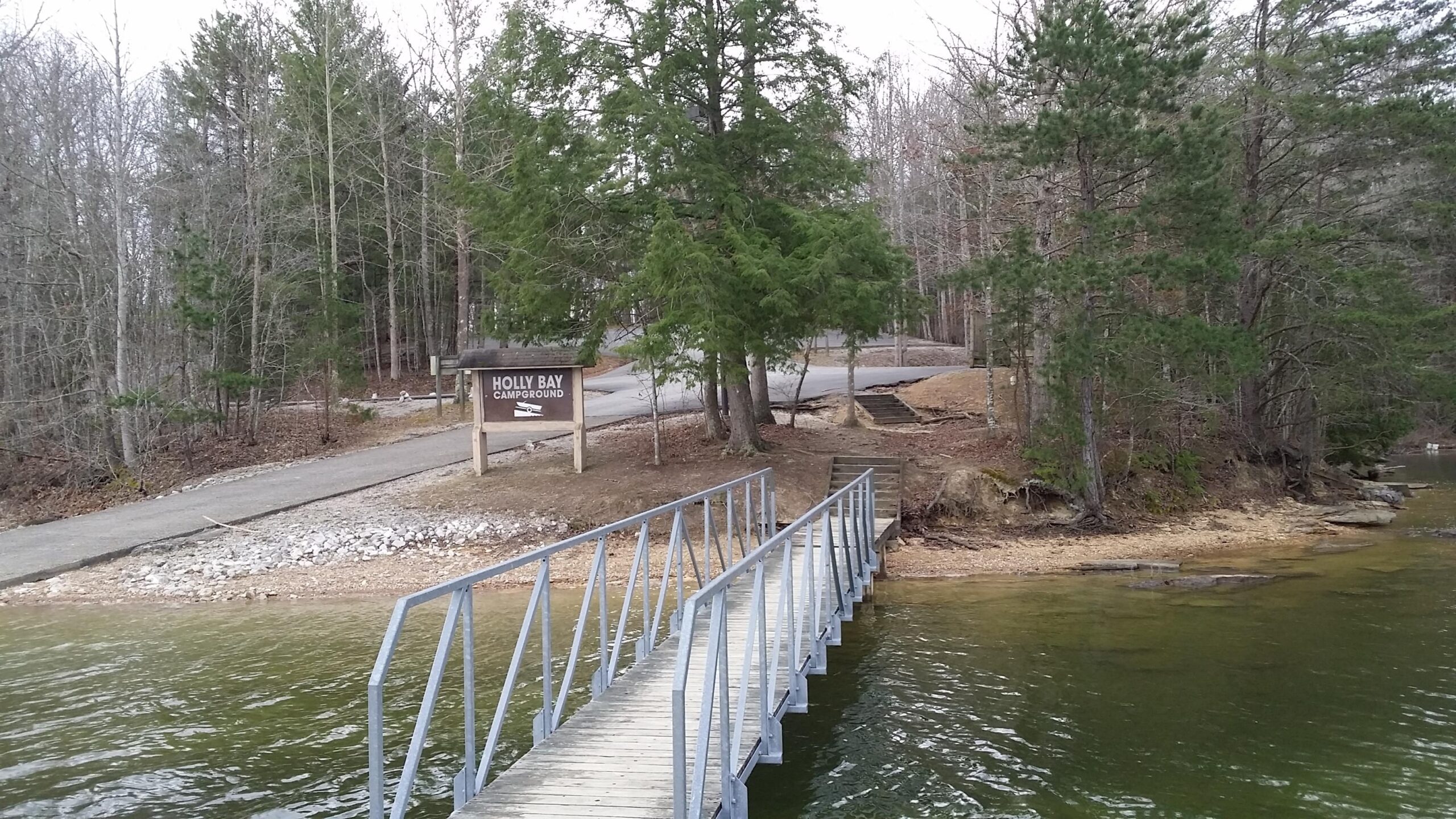 A wooden footbridge leads from a sandy beach area to a sign that reads "Holly Bay Campground." Surrounding the sign are trees with sparse foliage, indicating early spring or late fall. A paved road is visible in the background, winding through the forested landscape. The water in the foreground is calm, reflecting the surroundings. Sheltowee Trace - Laurel Lake Trail mountain bike trail.