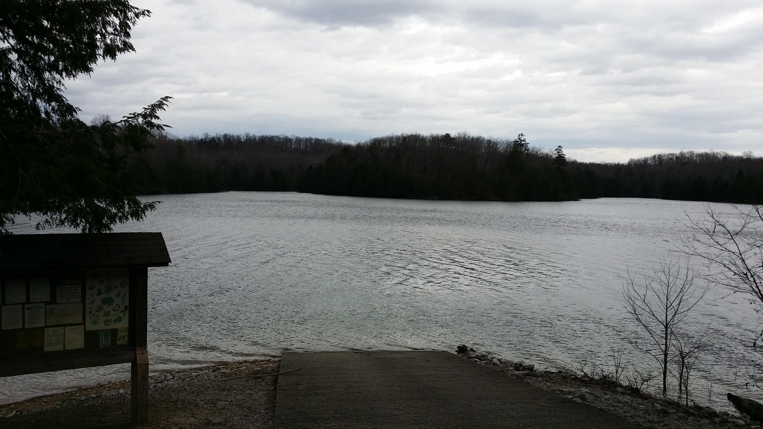 A tranquil view of a lake surrounded by trees under a cloudy sky. In the foreground, there is a wooden signboard with informational postings, along with a concrete pathway leading to the water's edge. The lake reflects the overcast sky, and the surrounding landscape is a mix of greenery and bare trees. Sheltowee Trace - Laurel Lake Trail mountain bike trail.