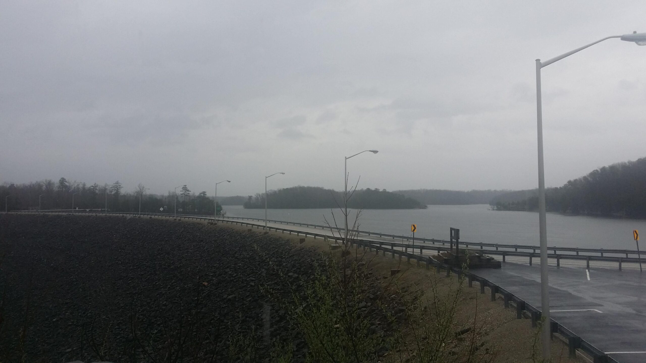 A view of a lake on a cloudy, rainy day, with a curved road partially visible in the foreground. Street lamps line the road, and an island can be seen in the distance across the water. The atmosphere is overcast and moody, suggesting inclement weather. Sheltowee Trace - Laurel Lake Trail mountain bike trail.