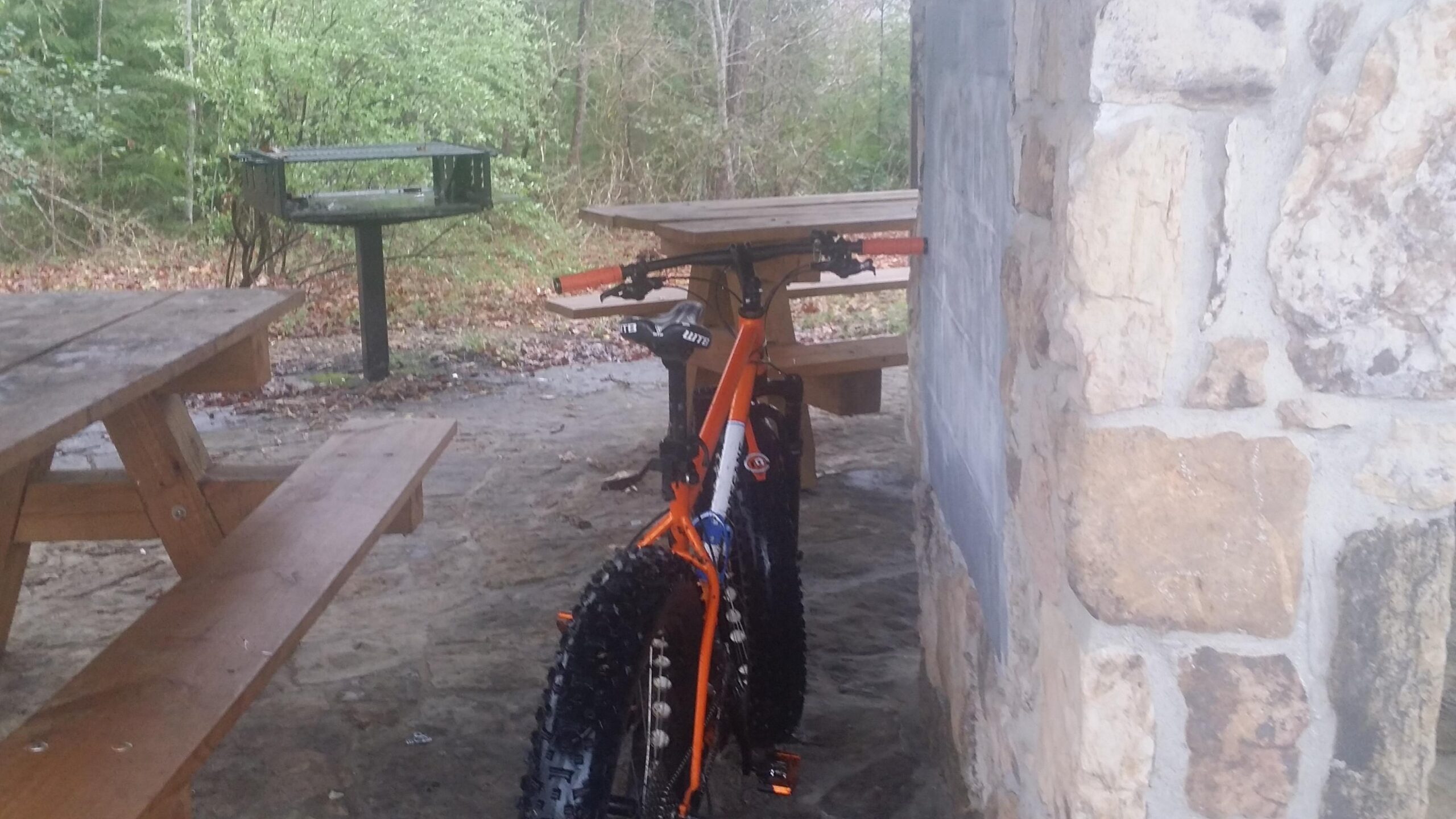 A vibrant orange mountain bike parked near a stone structure, with wooden picnic tables and a barbecue grill visible in the background. The setting is surrounded by green foliage, indicating an outdoor area. Sheltowee Trace - Laurel Lake Trail mountain bike trail.