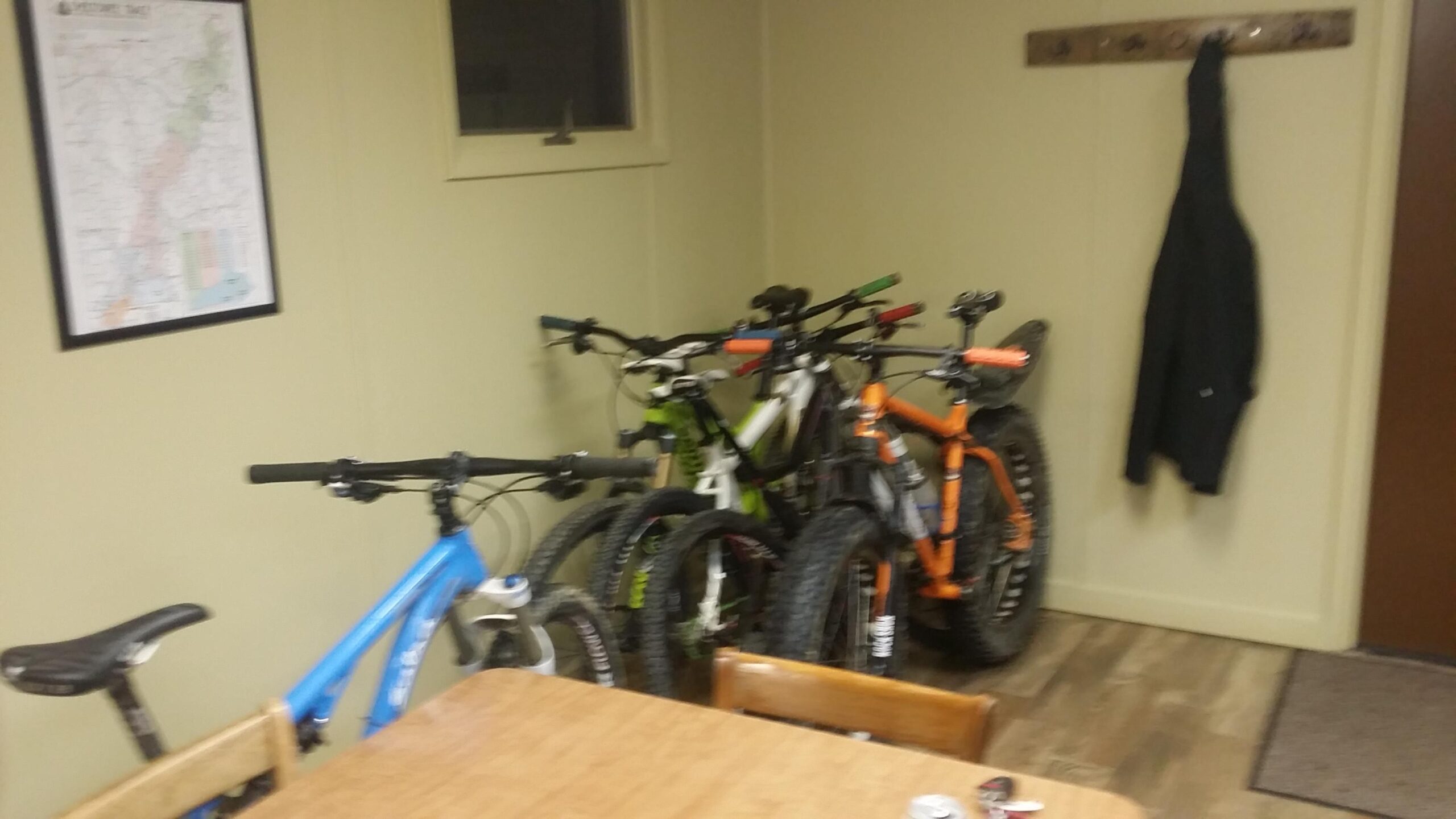 A cozy indoor space featuring a wooden table and several bicycles parked against the wall. The bikes include a blue mountain bike, a white bike, a green bike, and an orange bike with fat tires. A black jacket hangs on a hook nearby, and a map is framed on the wall. The flooring is wooden, creating a warm atmosphere. Sheltowee Trace - Laurel Lake Trail mountain bike trail.