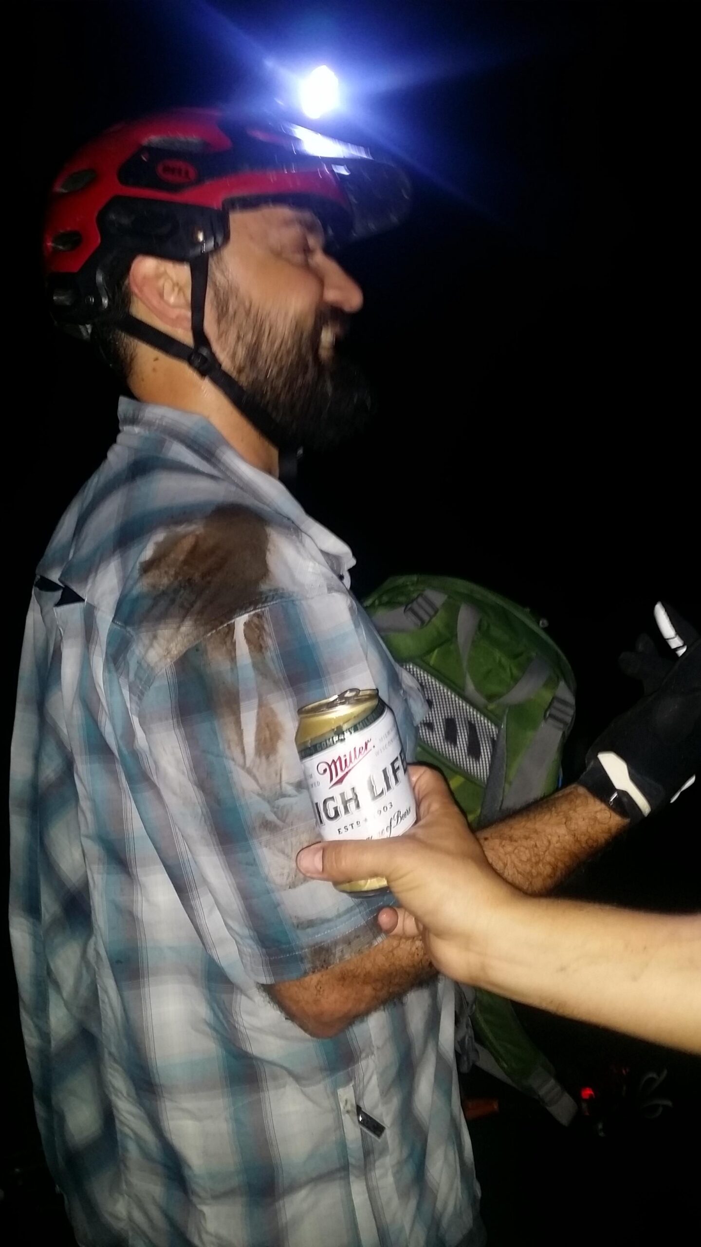 A man wearing a red helmet and a plaid shirt with a dirt mark on his shoulder smiles as he holds a can of Miller High Life. The background is dark, illuminated by a bright light source, possibly a headlamp. Another person's hand can be seen offering the can. A green backpack is visible in the background. Sheltowee Trace - Laurel Lake Trail mountain bike trail.