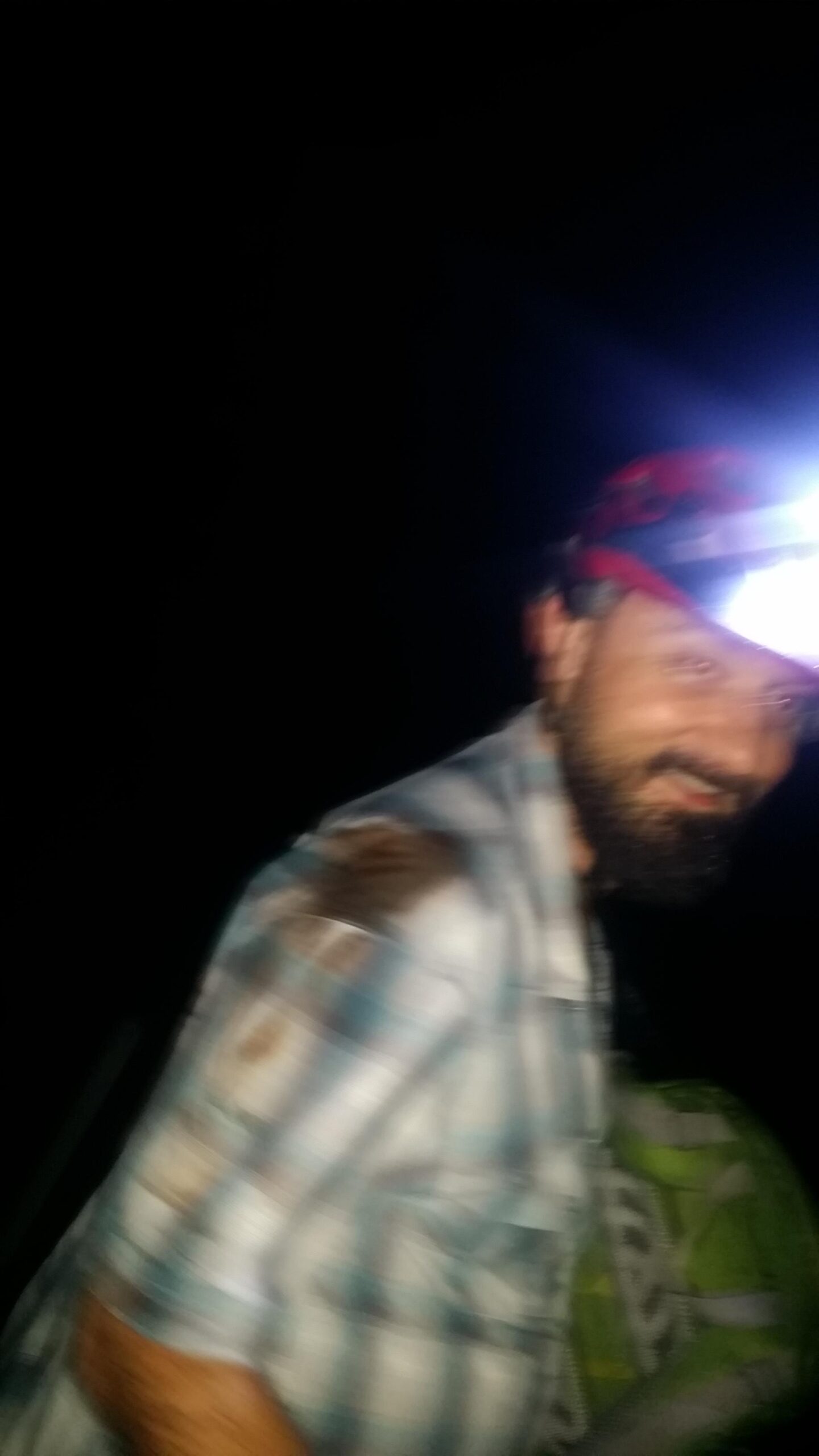 A slightly blurred image of a man wearing a checkered shirt and a headlamp, looking towards the camera against a dark background. He appears to be in a natural outdoor setting, possibly hiking or camping at night. Sheltowee Trace - Laurel Lake Trail mountain bike trail.
