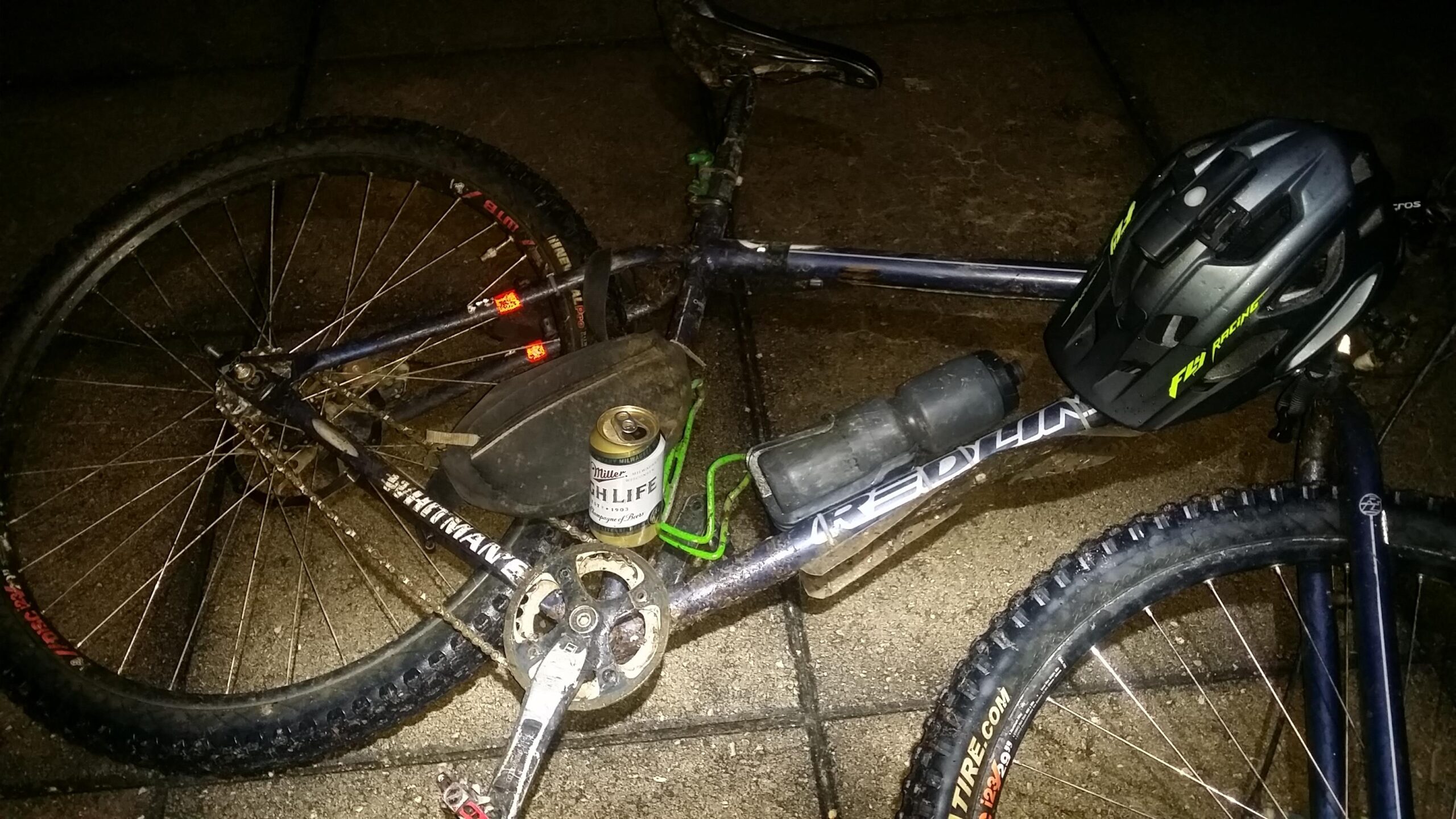 A close-up view of a mountain bike lying on a concrete surface at night, featuring a can of Miller High Life beer on the frame, a black helmet resting nearby, and a water bottle holder attached. The bike shows signs of use, with dirt on the tires and frame. Rear lights are visible on the bike, adding a splash of color in the dim lighting. Sheltowee Trace - Laurel Lake Trail mountain bike trail.