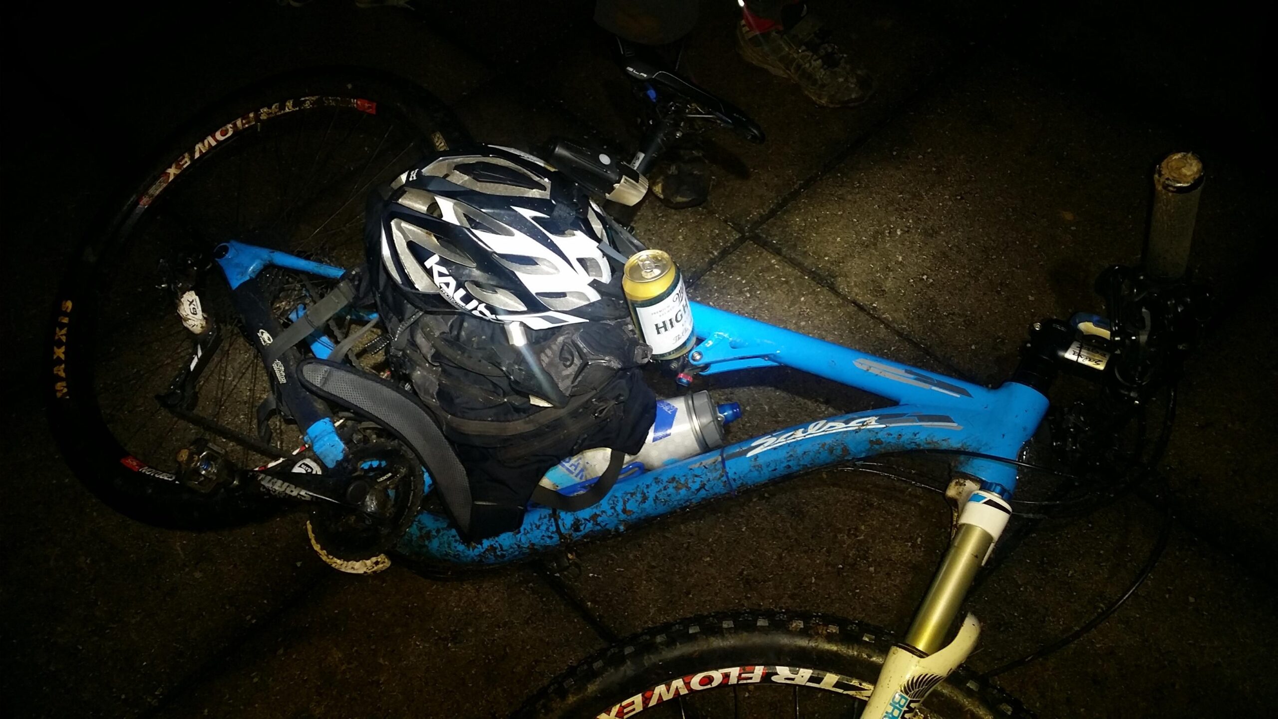 An illuminated blue mountain bike on a dark surface, featuring a black helmet and a backpack on its frame. A can of beer is resting on the bike alongside a water bottle. The bike's tires and frame show signs of mud, indicating recent use on rugged terrain. Sheltowee Trace - Laurel Lake Trail mountain bike trail.