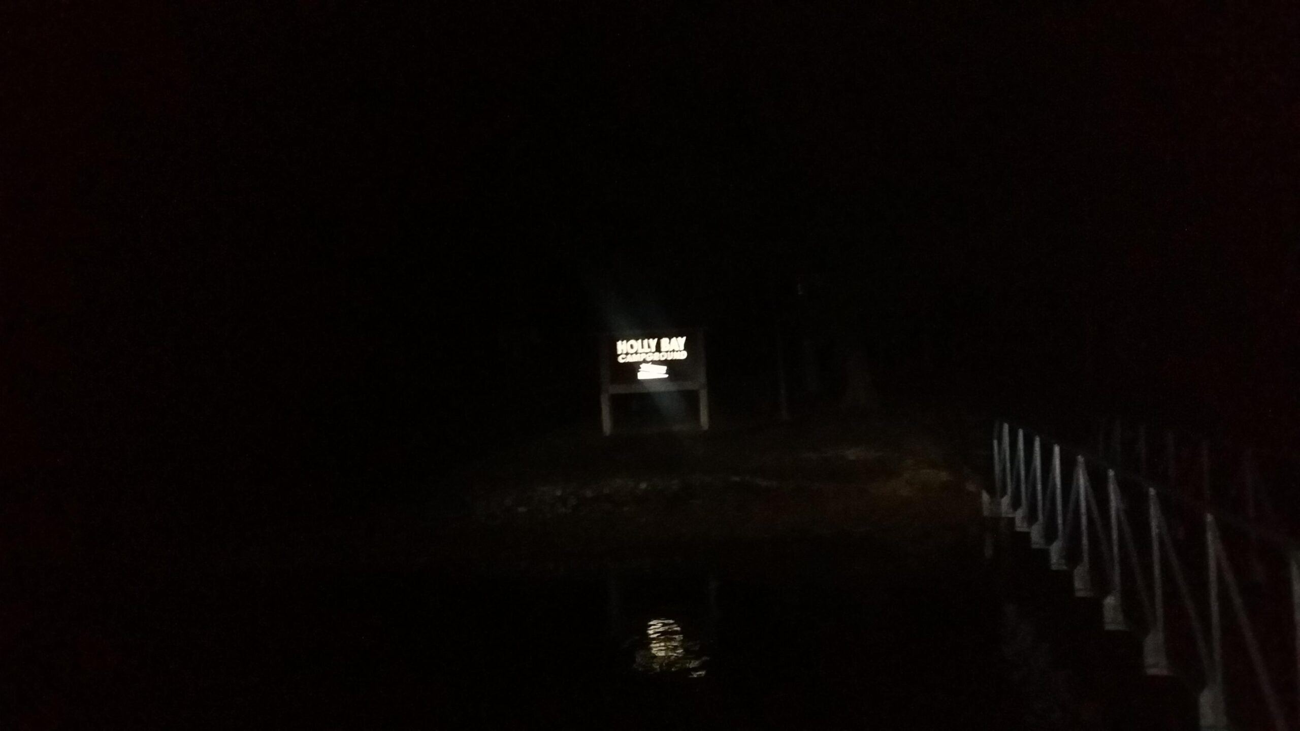 A dimly lit nighttime image of a sign that reads "Holly Bay Campground," partially illuminated by a light source. The background is dark, and there is a faint reflection visible on the water, along with a bridge made of metal railing in the foreground. Sheltowee Trace - Laurel Lake Trail mountain bike trail.