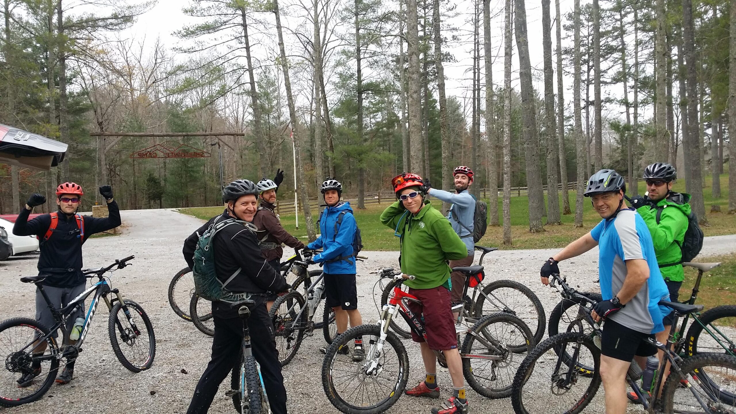 A group of eight mountain bikers, wearing helmets and cycling attire, standing with their bikes in a forested area. They are smiling and posing for the photo, with some making hand gestures. In the background, there is a sign indicating a trail entrance, surrounded by trees and a gravel path. Sheltowee Trace - Laurel Lake Trail mountain bike trail.