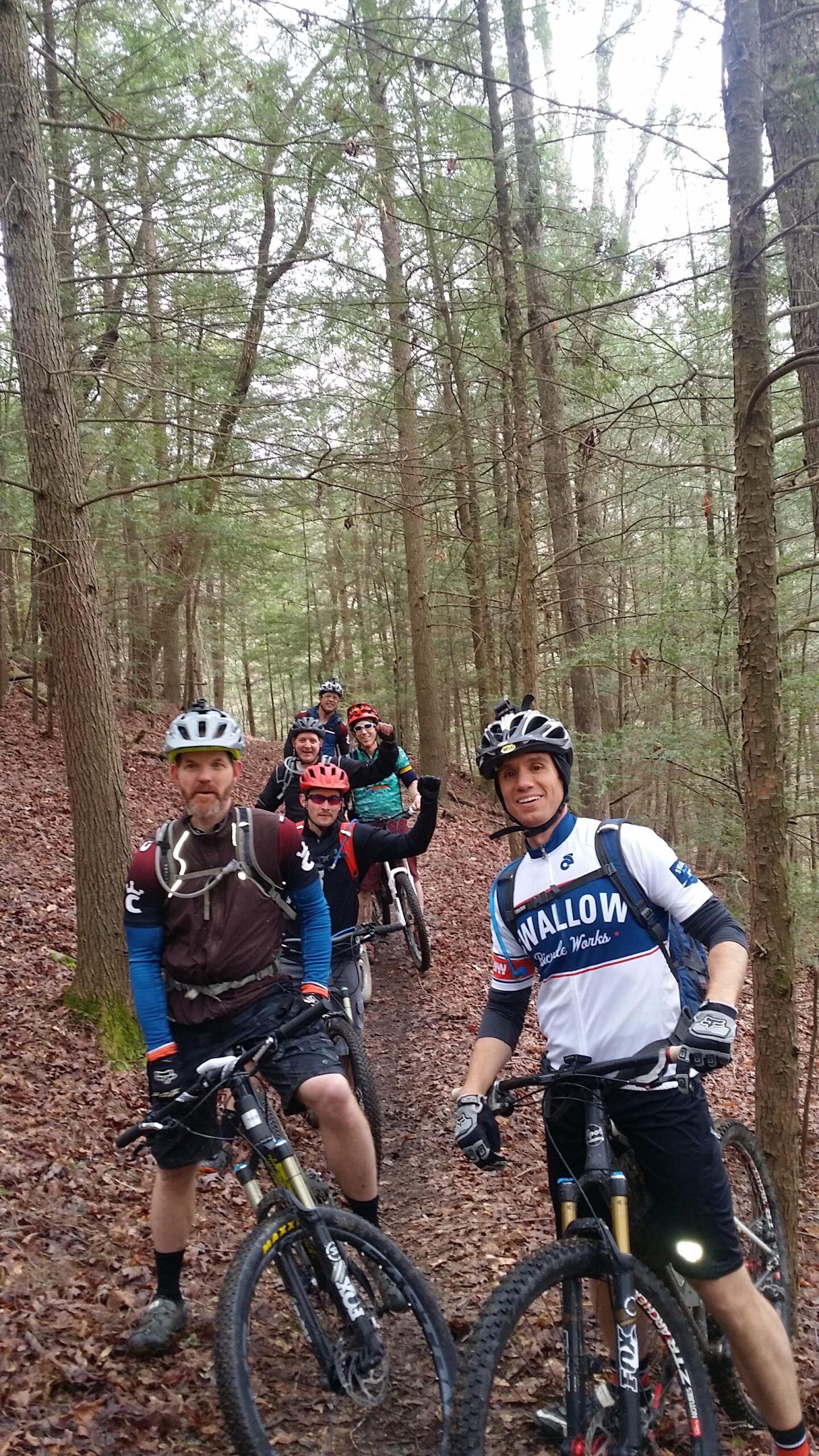 A group of six mountain bikers pause on a wooded trail, surrounded by tall trees and fallen leaves. They are wearing helmets and cycling gear, and some are smiling and posing for the camera while holding their bikes. The scene captures the camaraderie of outdoor biking in a natural setting. Sheltowee Trace - Laurel Lake Trail mountain bike trail.