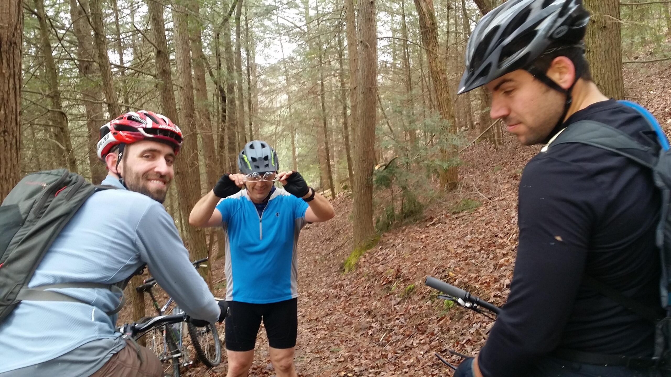Three mountain bikers pause on a wooded trail, surrounded by trees and fallen leaves. One cyclist, wearing a red helmet and gray shirt, smiles at the camera. Another, dressed in a blue shirt and black shorts, adjusts his sunglasses while standing in the foreground. The third cyclist, in a black shirt with a backpack, looks slightly away. Their bicycles are parked nearby, and the atmosphere is casual and friendly. Sheltowee Trace - Laurel Lake Trail mountain bike trail.