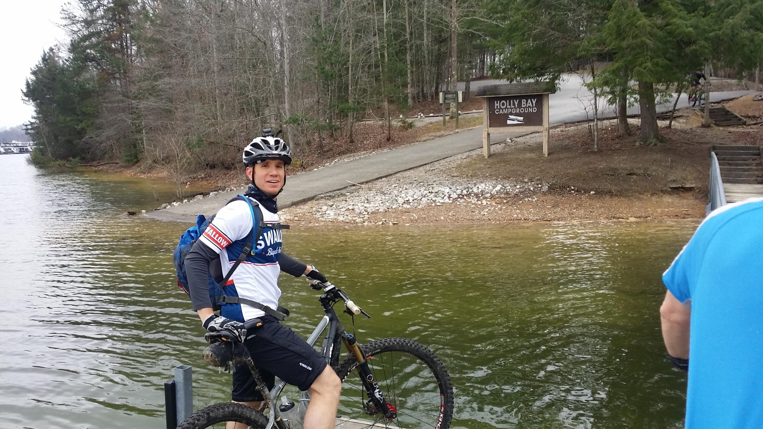 A person wearing a helmet and cycling gear is standing on the edge of a lake next to a mountain bike. In the background, there is a sign for "Holly Bay Campground," with a pathway leading into the woods. The scene features a calm water surface surrounded by trees and a pebbly shore, indicating a recreational area. Sheltowee Trace - Laurel Lake Trail mountain bike trail.
