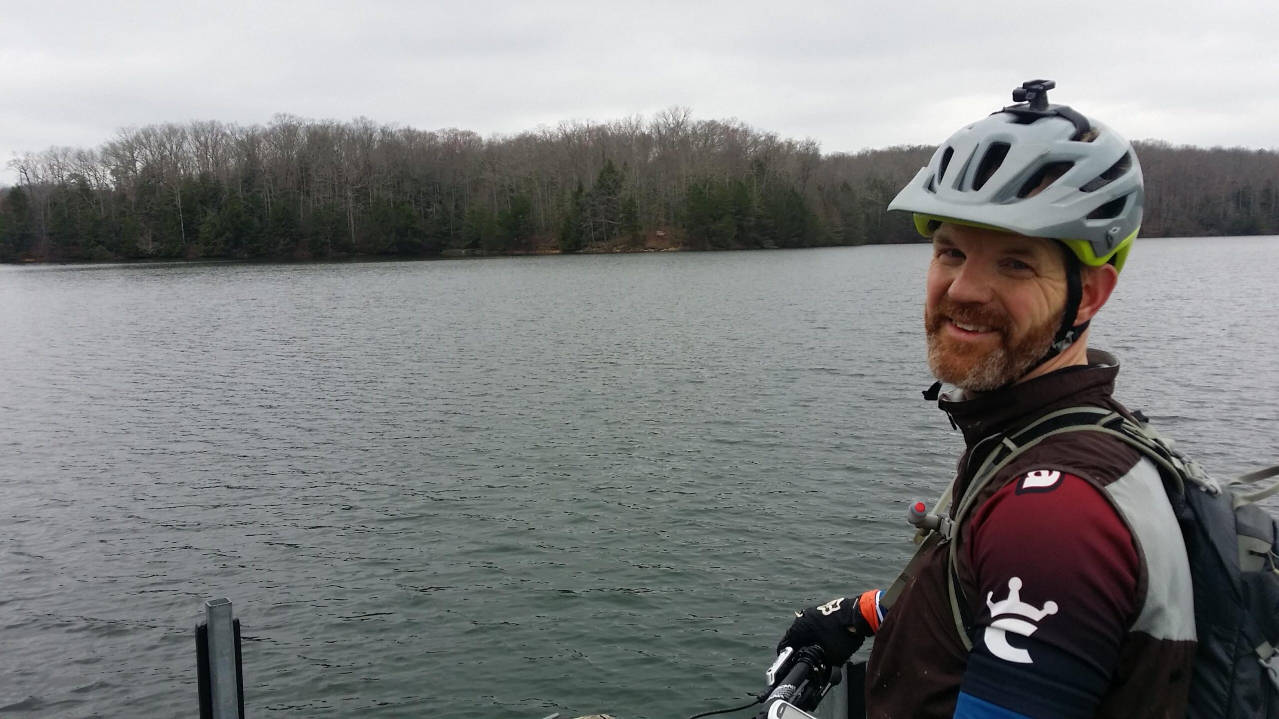 A smiling man wearing a bicycle helmet and cycling gear stands by a body of water, with trees in the background on a cloudy day. He leans on his mountain bike, enjoying the view. Sheltowee Trace - Laurel Lake Trail mountain bike trail.