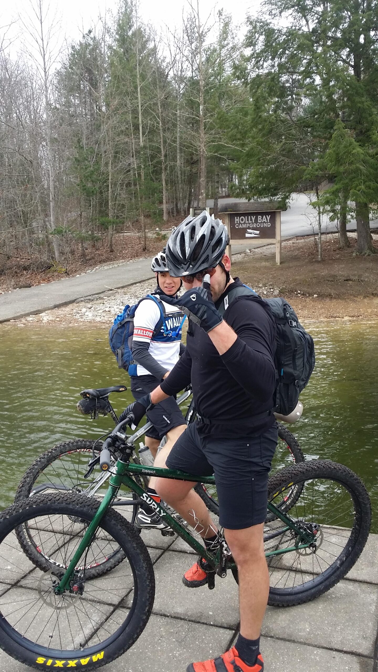 Two mountain bikers are posing near a body of water, with one biker wearing a dark outfit and red shoes, giving a thumbs-up gesture. The second biker, in a white shirt and backpack, smiles in the background. They are surrounded by trees, and a sign for "Holly Bay Campground" is visible nearby. The scene appears to be during an overcast day, suggesting an outdoor adventure. Sheltowee Trace - Laurel Lake Trail mountain bike trail.
