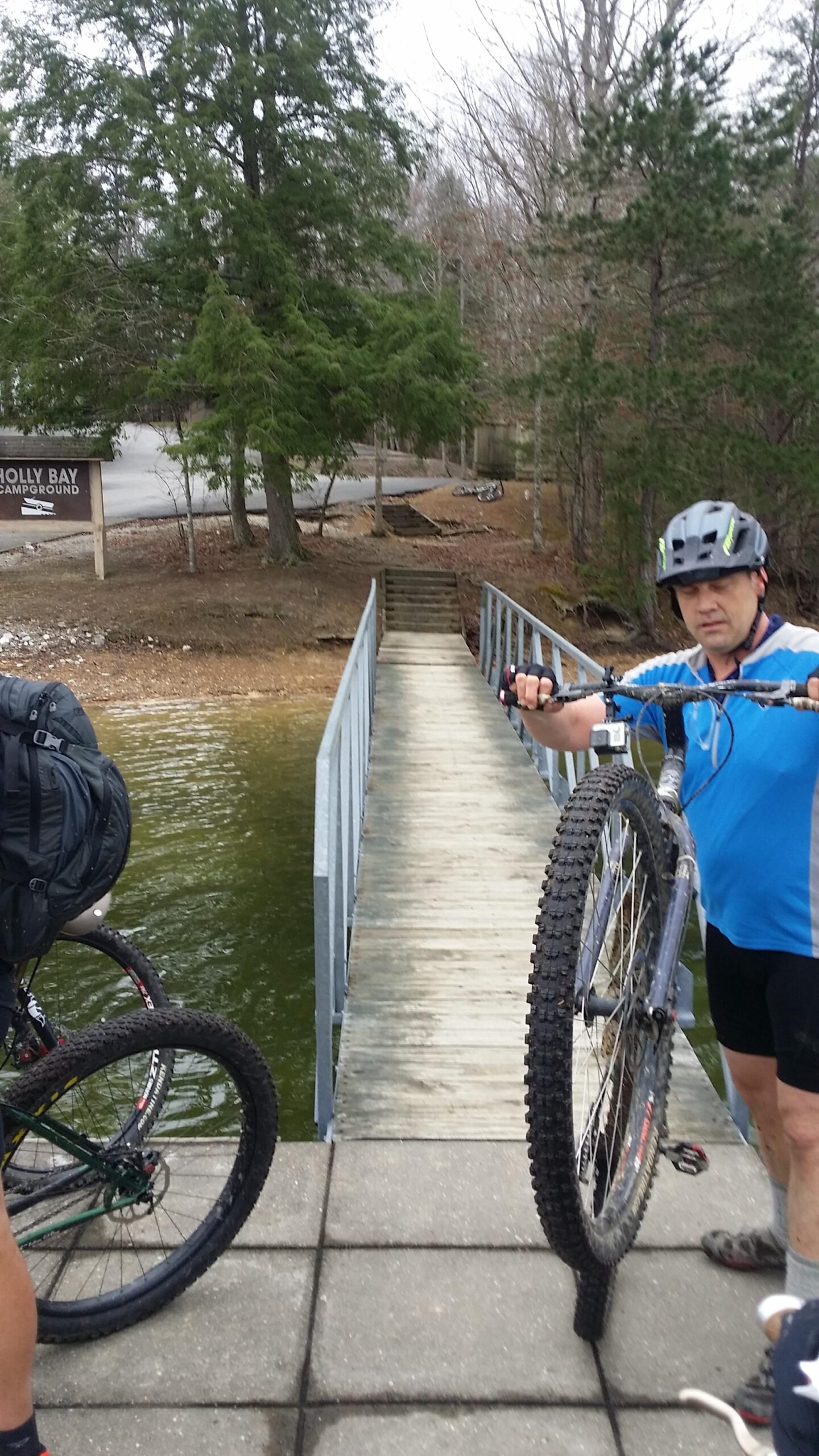A cyclist in a blue shirt holds a mountain bike wheel while standing on a wooden dock at Holly Bay Campground, with water visible on one side and a wooded area in the background. Another bicycle is nearby, and the scene is set in a natural, outdoor environment. Sheltowee Trace - Laurel Lake Trail mountain bike trail.