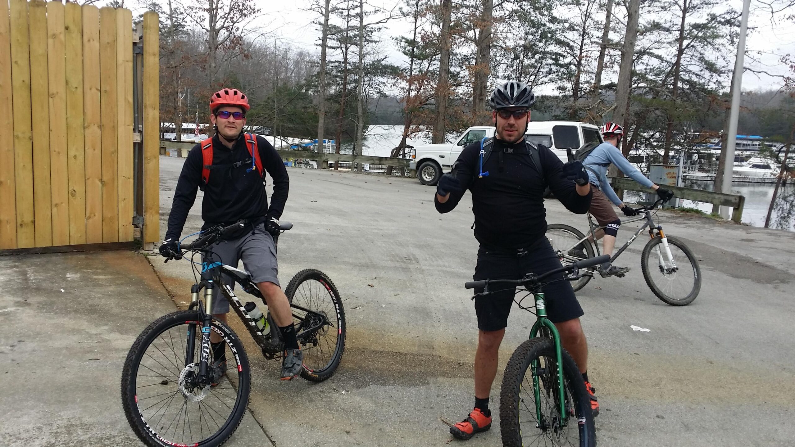 Two mountain bikers pose with their bikes in a gravel parking area near a wooden gate. One rider, wearing a red helmet and a backpack, is leaning on his bike with a slight smile. The other, in a black helmet and shirt, gives a thumbs-up. In the background, another biker rides away, and a lake with boats is visible through the trees. The environment appears to be overcast, indicating cool weather. Sheltowee Trace - Laurel Lake Trail mountain bike trail.