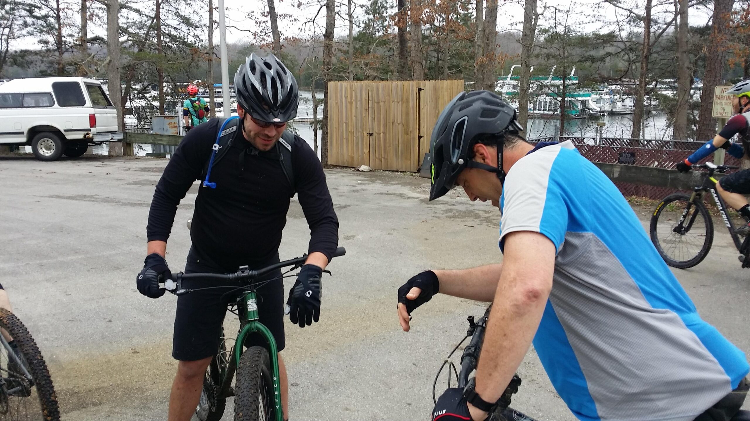 Two mountain bikers are adjusting their bikes in a parking area near a marina, with a white truck and watercraft in the background. One biker, dressed in a black shirt and gloves, is discussing the bike with another biker who is wearing a blue and gray shirt. They appear focused on their equipment. Sheltowee Trace - Laurel Lake Trail mountain bike trail.