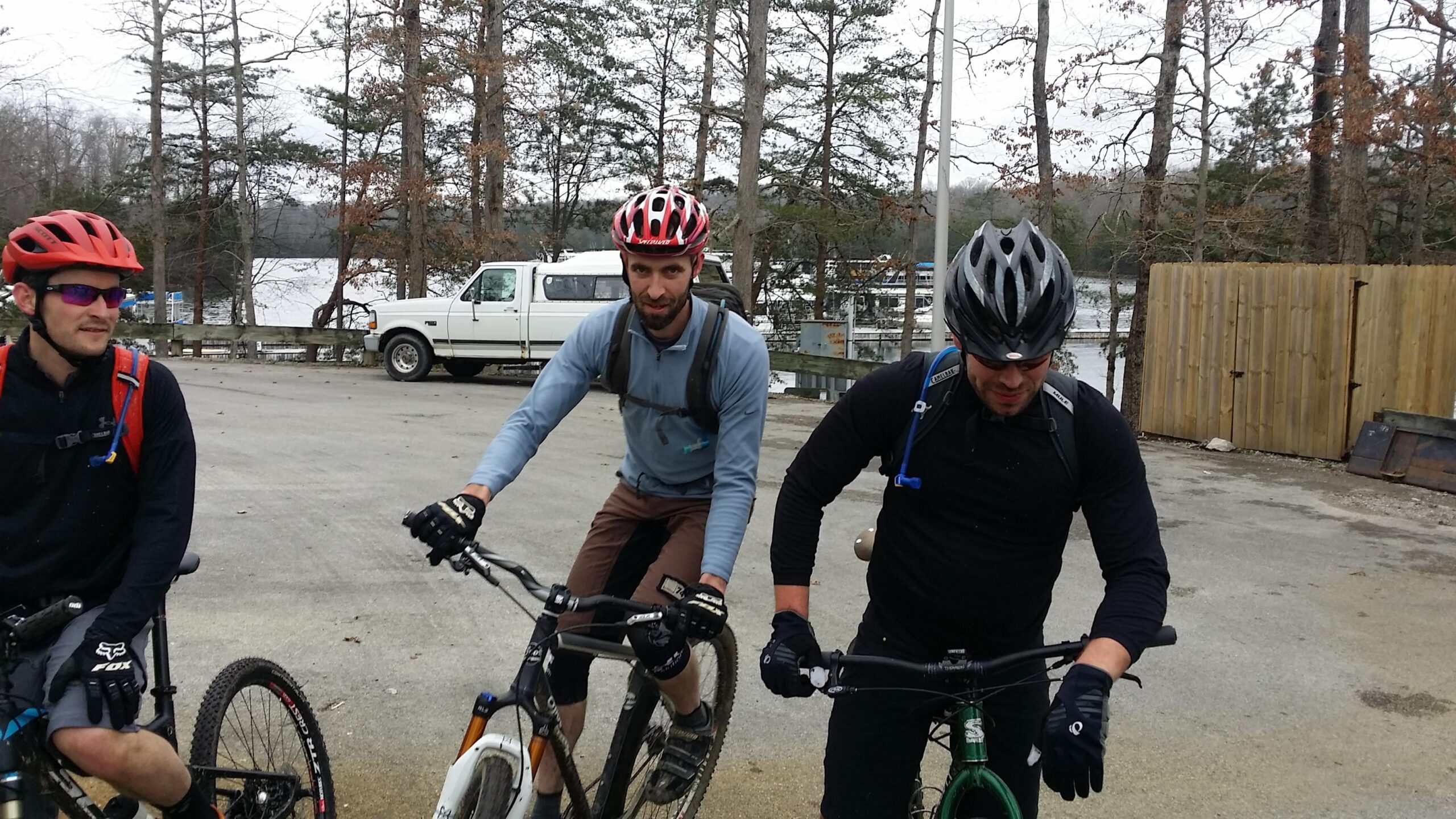 Three mountain bikers are gathered by their bikes in a gravel parking area near a lake. The first rider, wearing a red helmet and sunglasses, is seated on his bike. The second rider, dressed in a blue long-sleeve shirt and brown pants, is adjusting his grip on the handlebars. The third rider, in a black long-sleeve shirt and gloves, is leaning forward on his green bike. In the background, trees line the lake, and a white truck is parked nearby. The weather appears overcast. Sheltowee Trace - Laurel Lake Trail mountain bike trail.