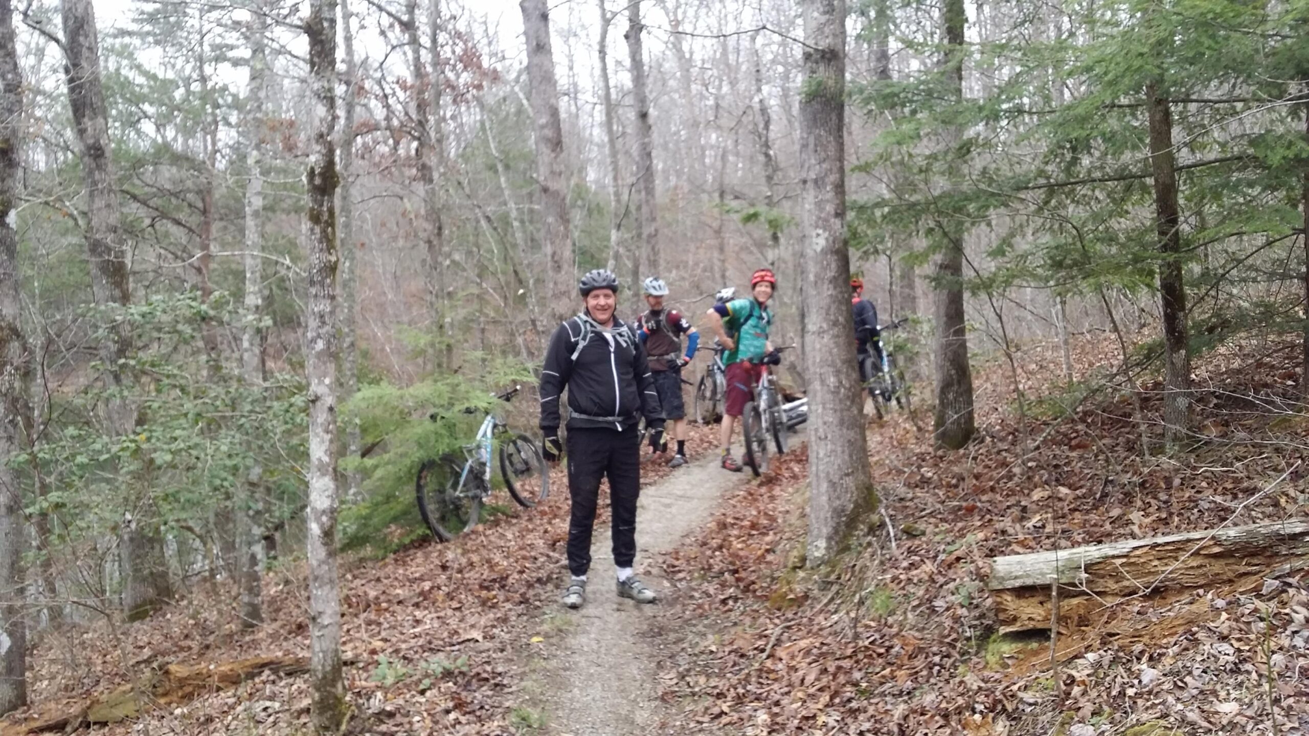 A group of six mountain bikers are standing on a dirt trail in a wooded area, surrounded by trees and fallen leaves. One biker, in the foreground, is smiling and wearing a black helmet, jacket, and cycling shoes. The others are positioned along the trail, some with their bikes, enjoying a break during their ride. The atmosphere appears relaxed and friendly, with a backdrop of a serene forest setting. Sheltowee Trace - Laurel Lake Trail mountain bike trail.