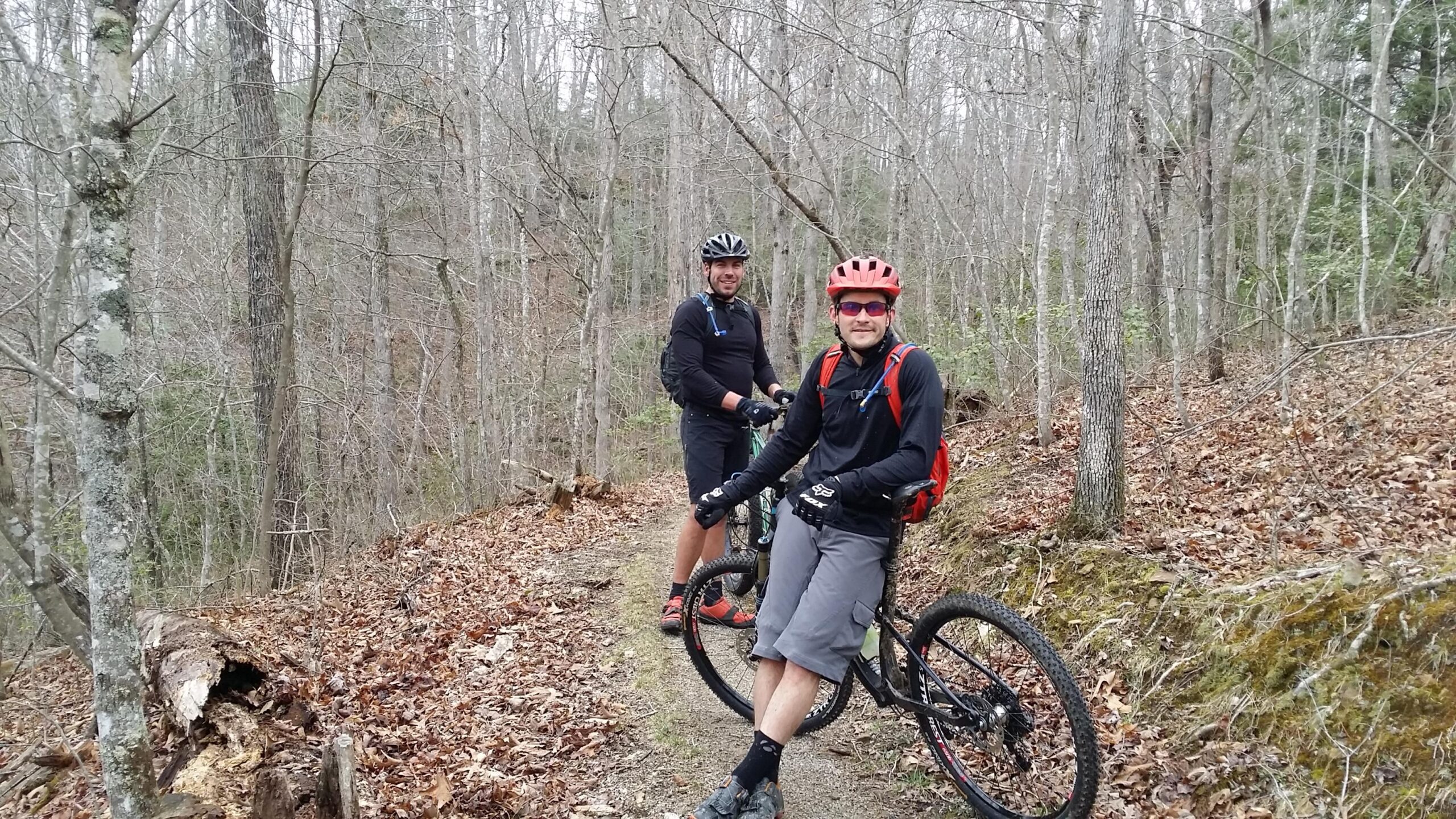 Two mountain bikers pause on a dirt trail surrounded by bare trees and scattered leaves. One biker wearing a red helmet and a black long-sleeve shirt leans against his bike, while the other, wearing a black helmet, stands nearby, both smiling. The scene conveys a moment of camaraderie in an outdoor setting. Sheltowee Trace - Laurel Lake Trail mountain bike trail.