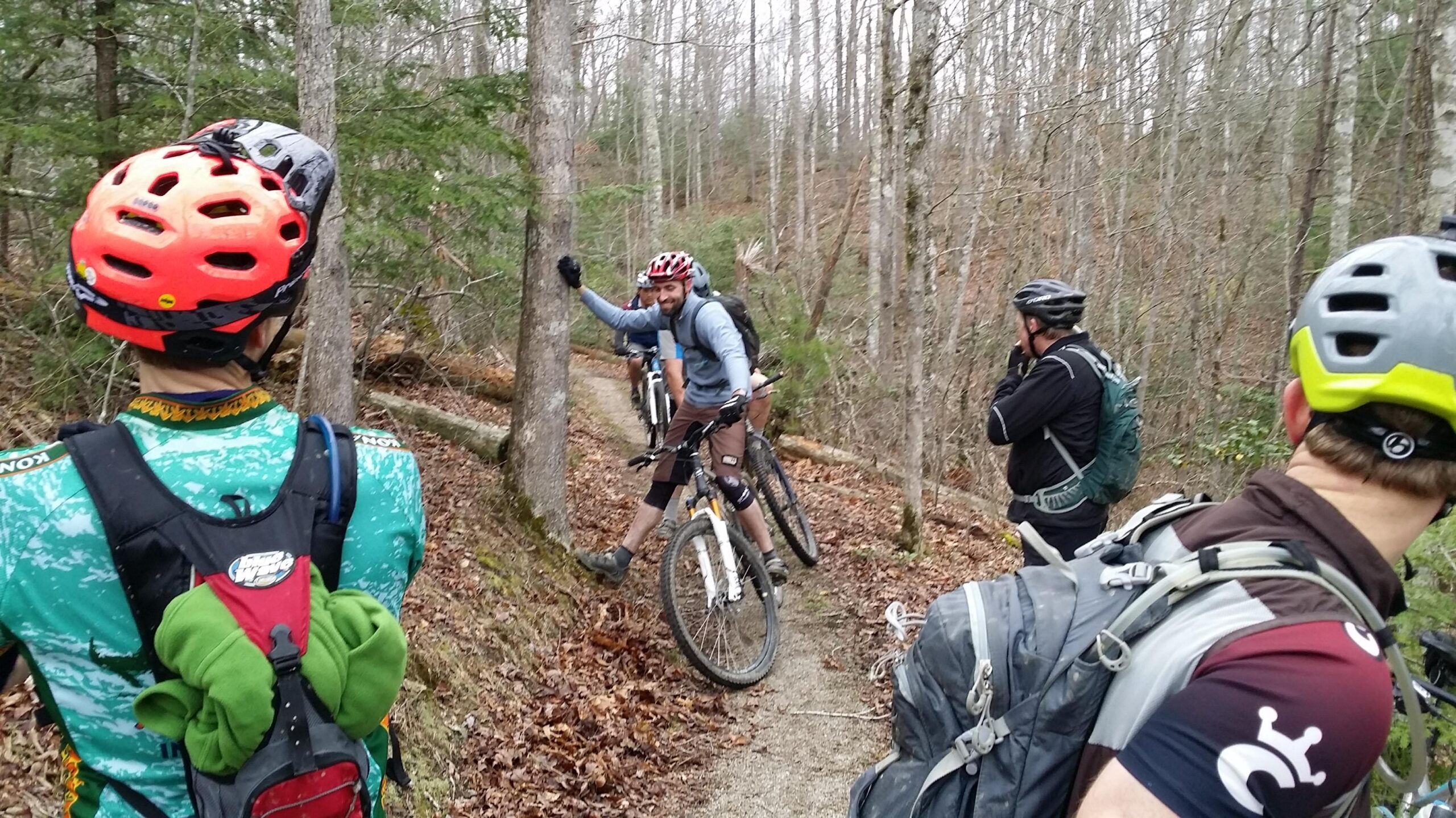 A group of mountain bikers stops on a wooded trail, discussing their route. One biker is leaning against a tree while others are positioned nearby, wearing helmets and riding gear. The landscape features bare trees and fallen leaves, indicating a cool season. Sheltowee Trace - Laurel Lake Trail mountain bike trail.