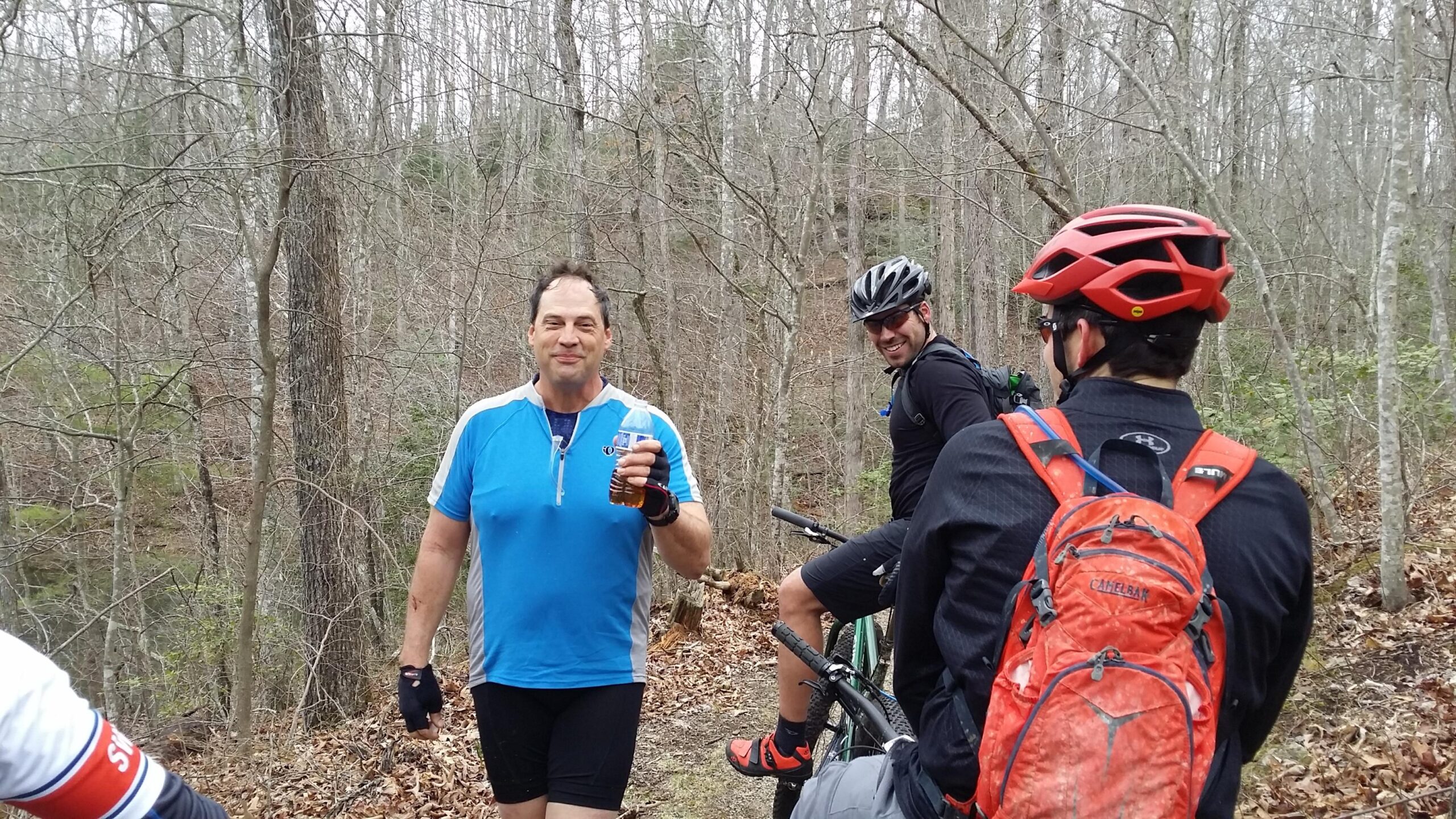 Three cyclists are on a wooded trail during an outdoor ride. One man, wearing a blue cycling shirt and holding a drink, is smiling and facing the camera. Behind him, another cyclist in black gear is seated on a bicycle, looking back and smiling. A third cyclist, mostly out of view, is wearing a red helmet and a backpack. The background features bare trees and dry leaves, suggesting an early spring setting. Sheltowee Trace - Laurel Lake Trail mountain bike trail.