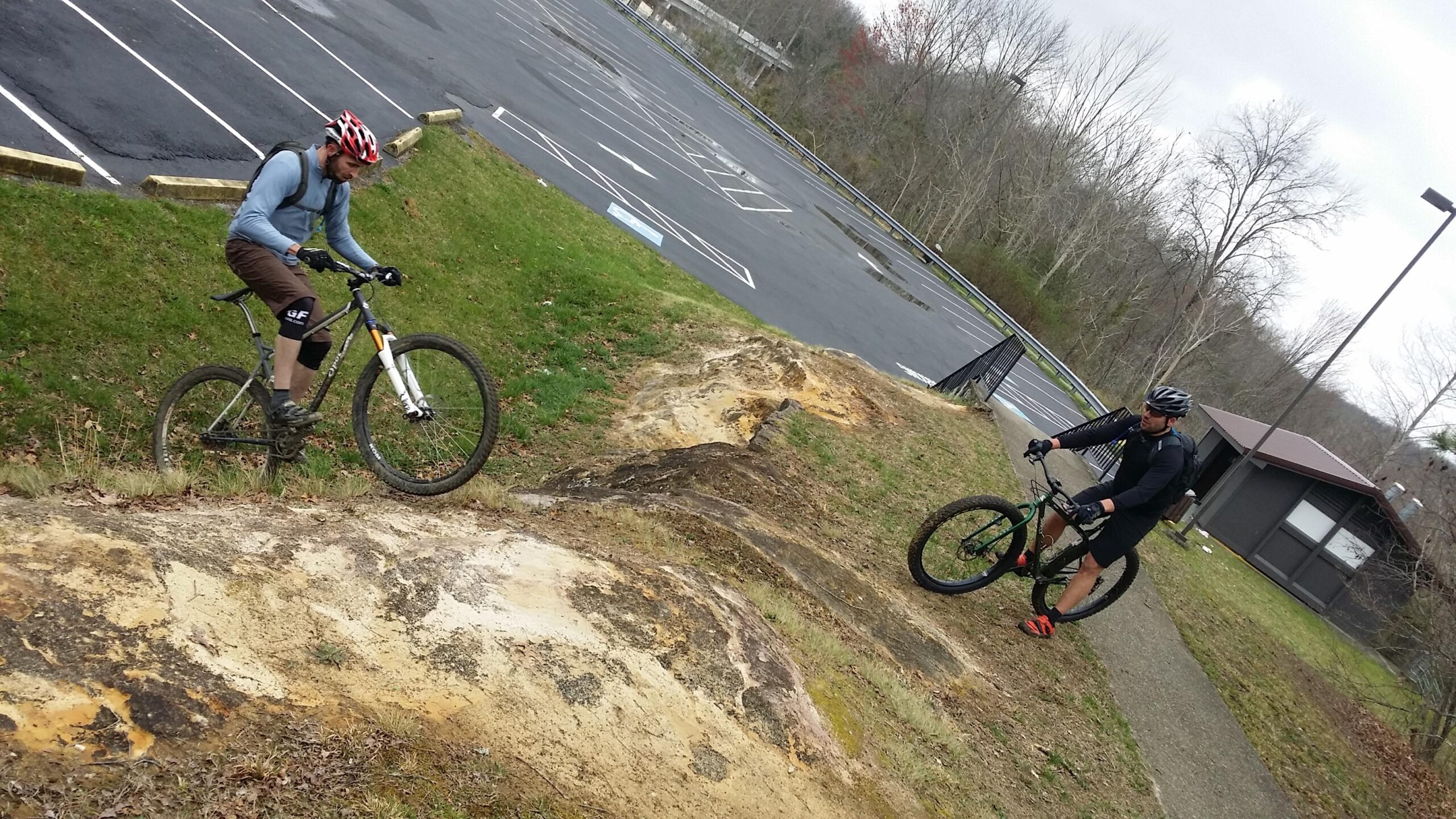 Two cyclists on a grassy hillside with a dirt path. One rider is navigating a steep incline on a mountain bike, while the other is standing beside theirs, wearing a helmet and athletic gear. The background shows an empty parking lot and some trees, with cloudy skies overhead. Sheltowee Trace - Laurel Lake Trail mountain bike trail.