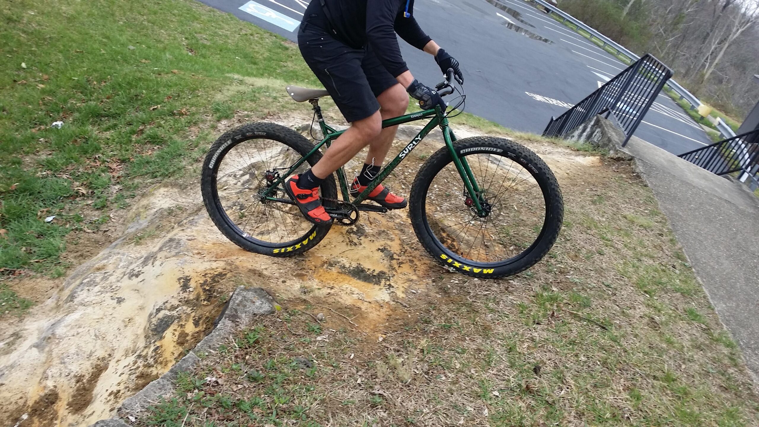 A person riding a green mountain bike with wide tires is navigating a dirt path. They are wearing black shorts and red cycling shoes, showing dynamic movement as they balance on a slope near a grassy area. In the background, a parking lot and a set of stairs are visible. Sheltowee Trace - Laurel Lake Trail mountain bike trail.