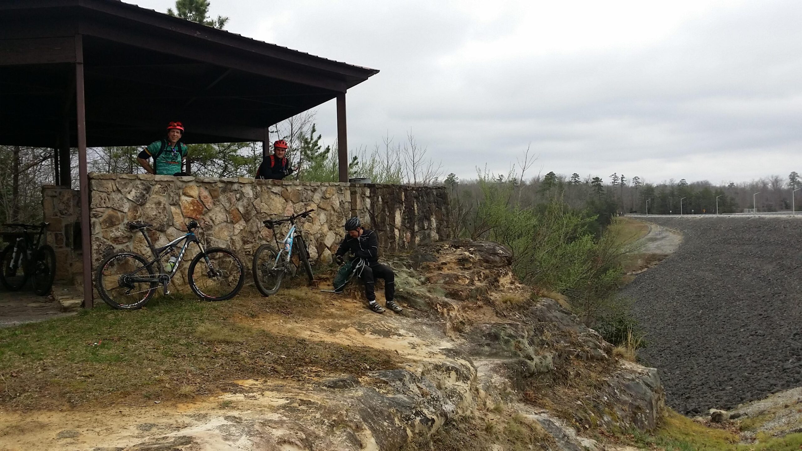 Three mountain bikers taking a break at a stone shelter beside a reservoir. Two cyclists are standing near their bikes, while a third sits on a rock, looking at a bag. The landscape features trees in the background and a gravel path leading away from the shelter. The sky is overcast, suggesting a cool day. Sheltowee Trace - Laurel Lake Trail mountain bike trail.