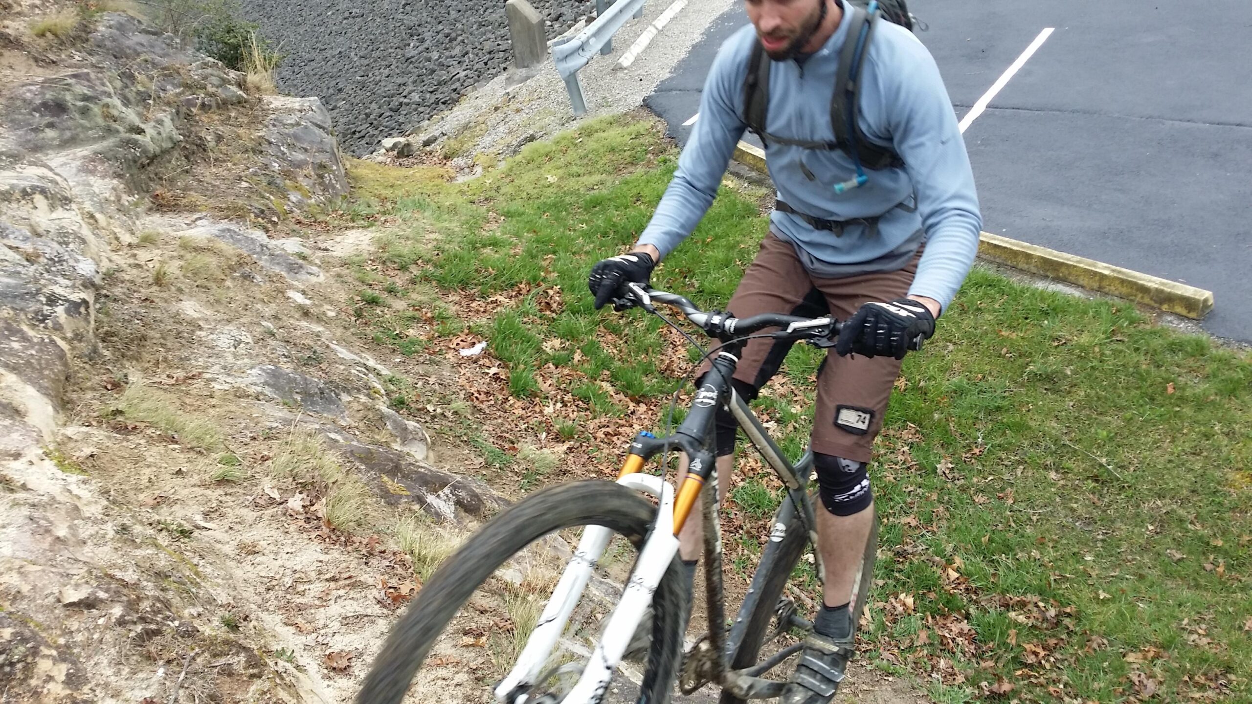 A mountain biker navigating a rocky and uneven terrain, using his bike to climb up a slope. The rider is wearing protective gear, including gloves and knee pads, and is focused on maintaining balance as he ascends. In the background, there are patches of grass and a gravel area indicating an outdoor cycling environment. Sheltowee Trace - Laurel Lake Trail mountain bike trail.