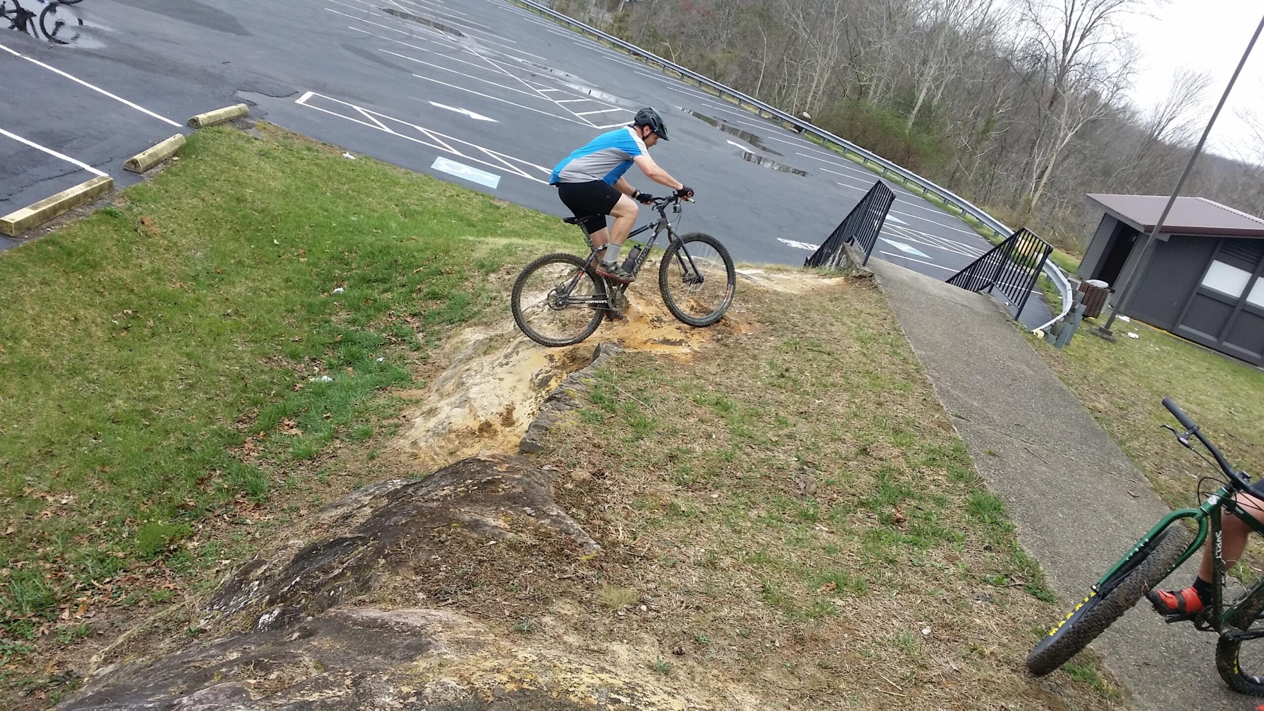 A cyclist in a blue and gray jersey is riding down a rocky incline on a mountain bike, navigating through a grassy area. In the background, a parking lot is visible, along with a building and a sloped pathway leading up to a railing. The scene captures an outdoor biking activity in a natural setting. Sheltowee Trace - Laurel Lake Trail mountain bike trail.