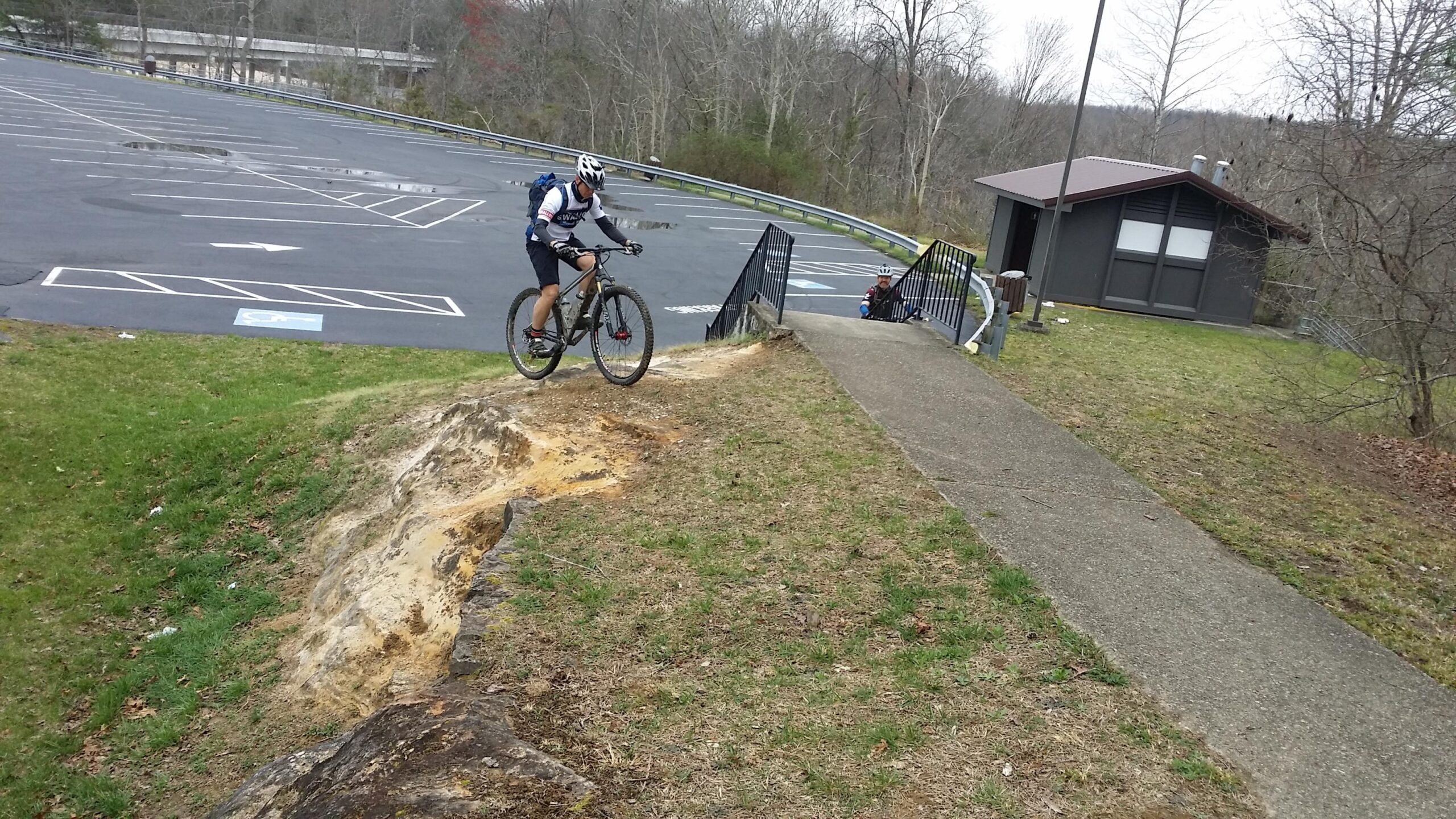 A mountain biker navigates a steep, sandy slope near a parking lot, while another person sits on steps nearby. The scene is set in a wooded area with bare trees in the background, and a small building is partially visible. Sheltowee Trace - Laurel Lake Trail mountain bike trail.