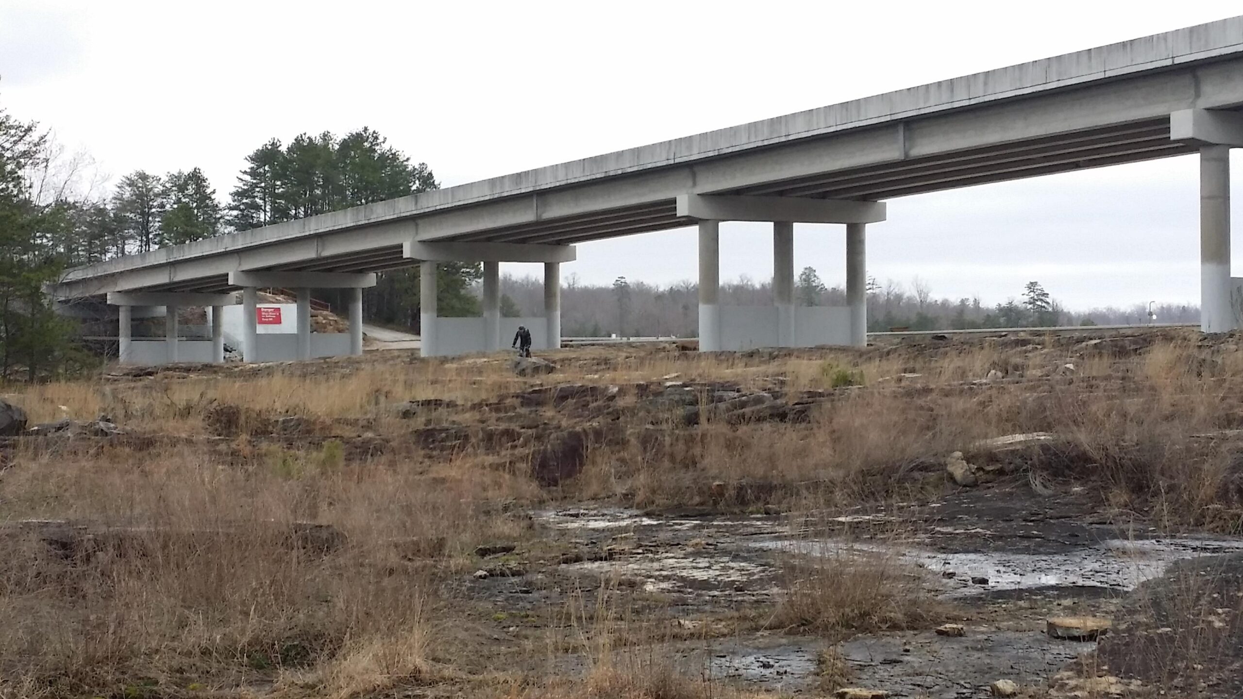 A view of an empty landscape featuring a concrete overpass supported by columns. The foreground is characterized by dry grass and rocky terrain, with a person walking beneath the bridge. In the background, sparse trees and a cloudy sky are visible. A sign is partially seen on the side of the overpass. Sheltowee Trace - Laurel Lake Trail mountain bike trail.