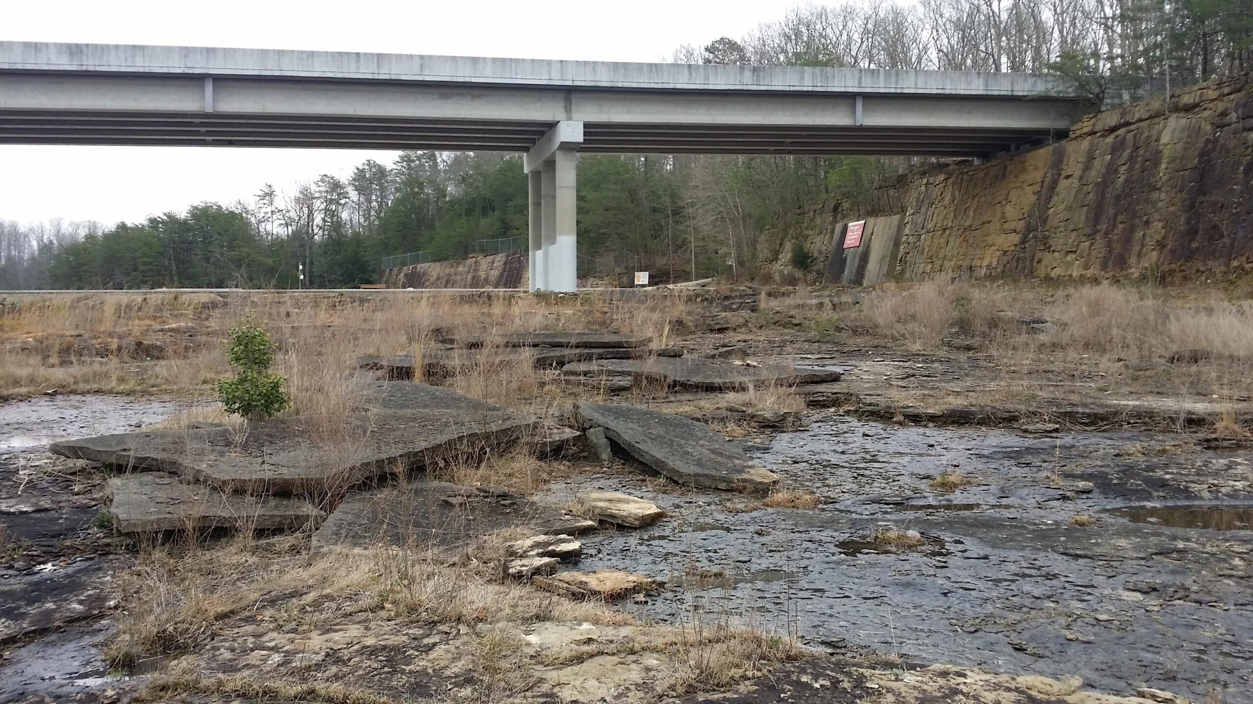 An overpass bridges a rocky landscape, where large stone slabs and patches of grass are visible. The area appears dry with sparse vegetation, and trees can be seen in the background. A sign on a wall next to the overpass is partially obscured. Sheltowee Trace - Laurel Lake Trail mountain bike trail.